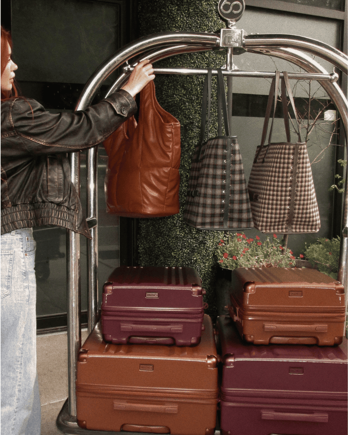 Woman hanging a CALPAK Luka Everything Tote in Vegan Leather Honey Brown on luggage trolley 