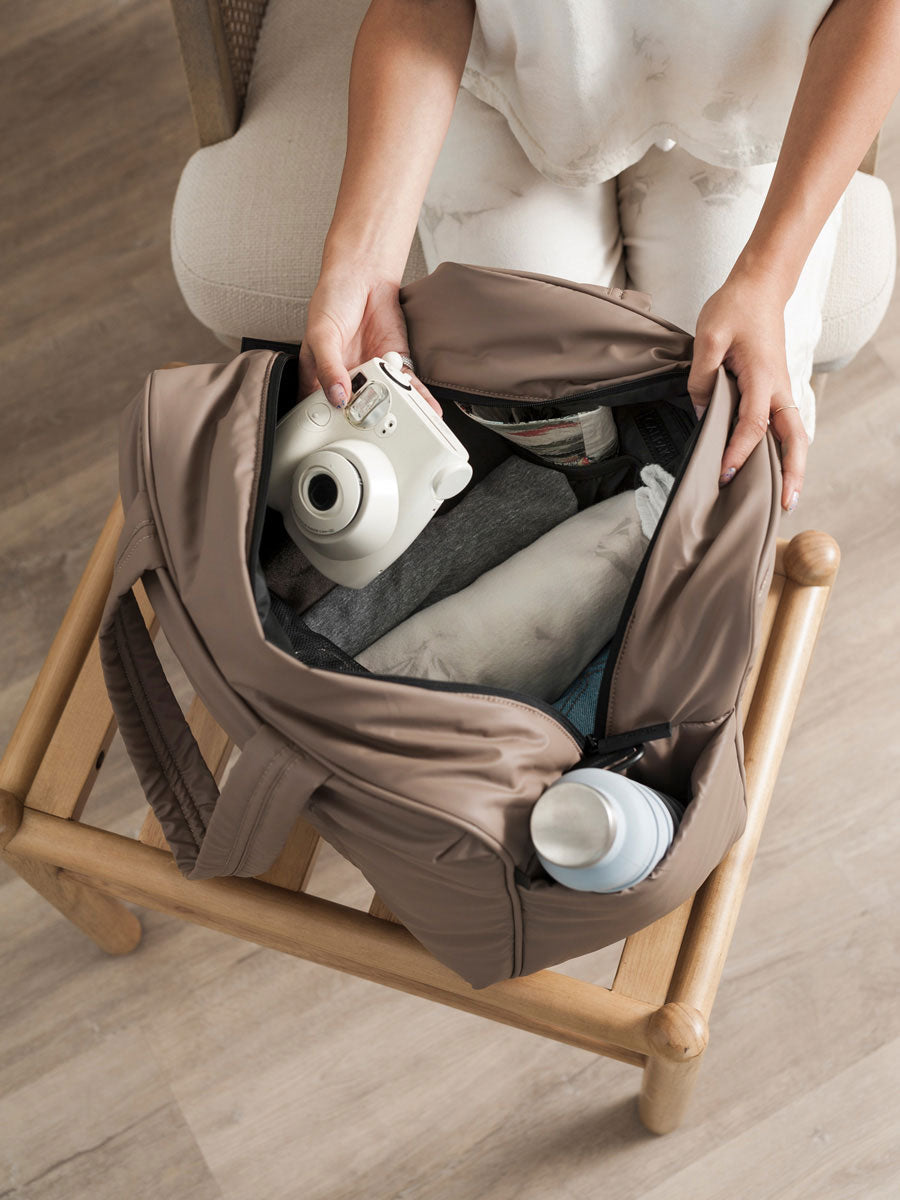 Person adding a polaroid and clothes into the CALPAK Luka Duffel in brown Chocolate