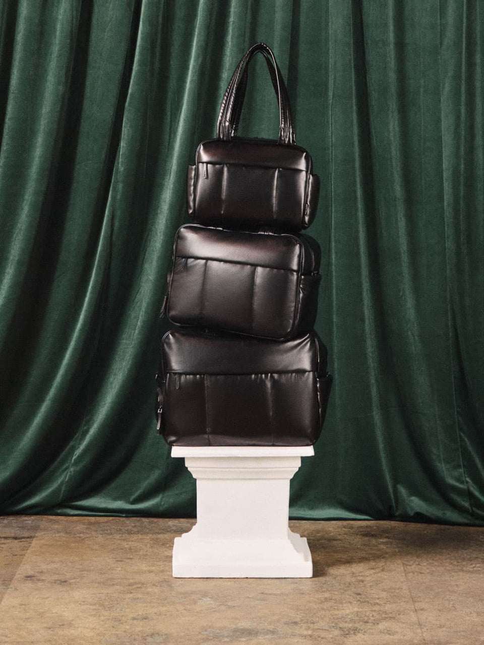 CALPAK Mini, Regular, and Large Luka Duffel Bags on a white pedestal against a green curtain background 