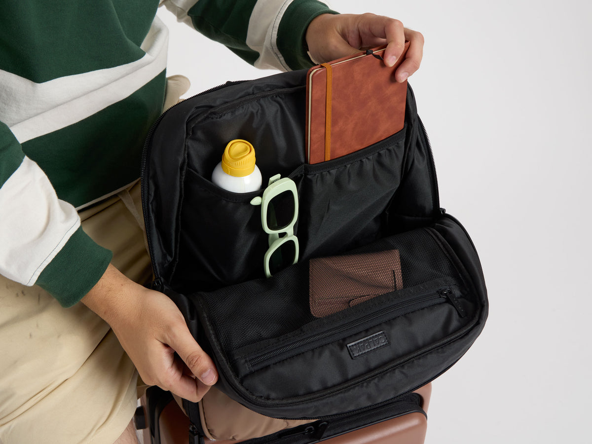 CALPAK Luka Laptop Backpack in brown Chocolate being opened by model to put in notebook and sunglasses into compartments