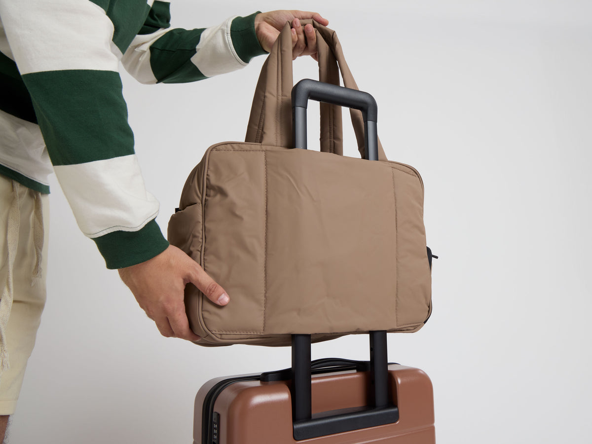 CALPAK Luka Duffel in brown Chocolate being put onto trolley of CALPAK Hue Carry On Luggage in brown Hazel by model 