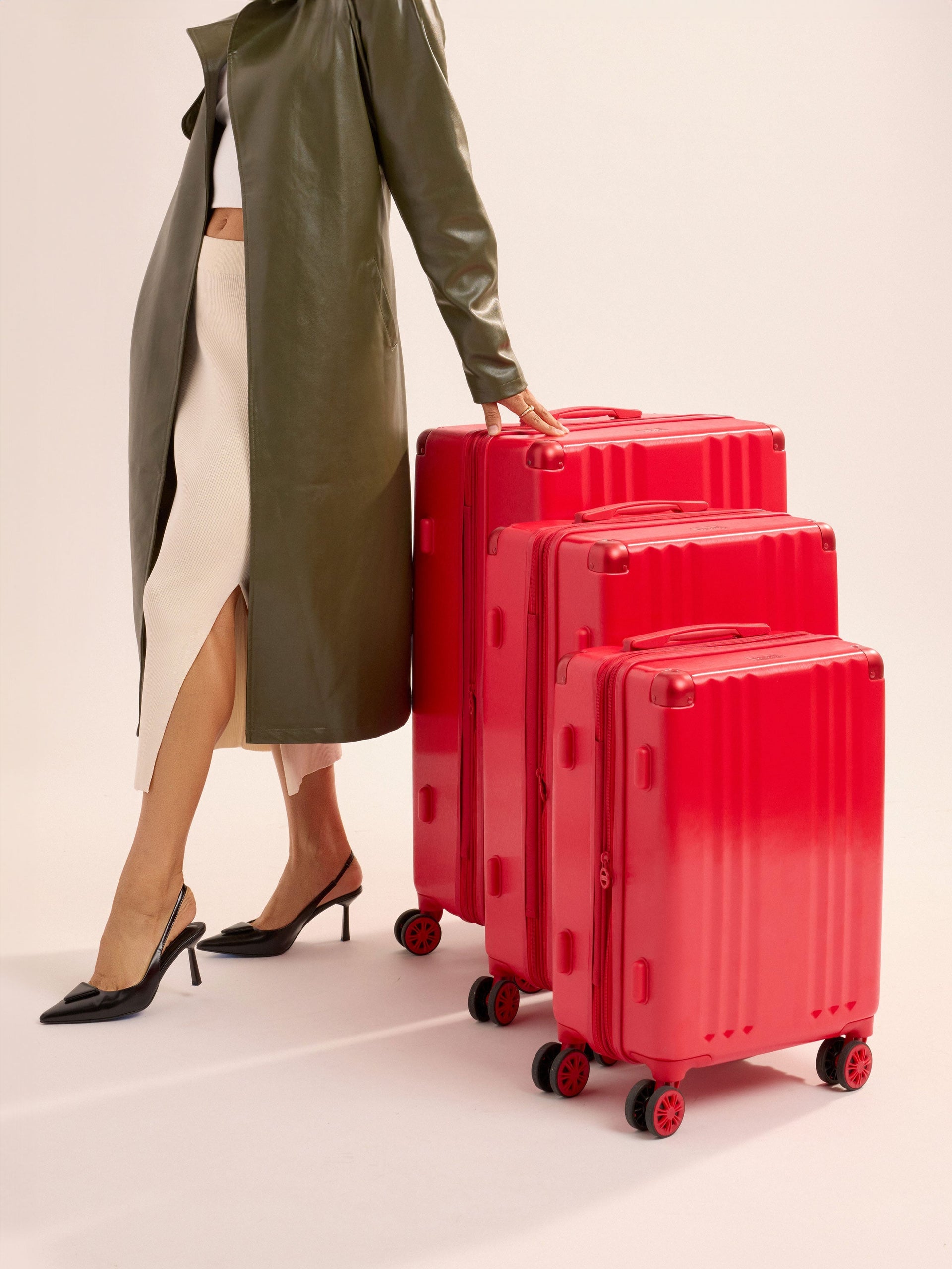 Model standing besides CALPAK Ambeur Large Luggage, Ambeur Medium Luggage and Ambeur Carry-On Luggage lined up tallest to shortest in metallic red, part of 3-piece luggage set; LAM3000-METALLIC-RED