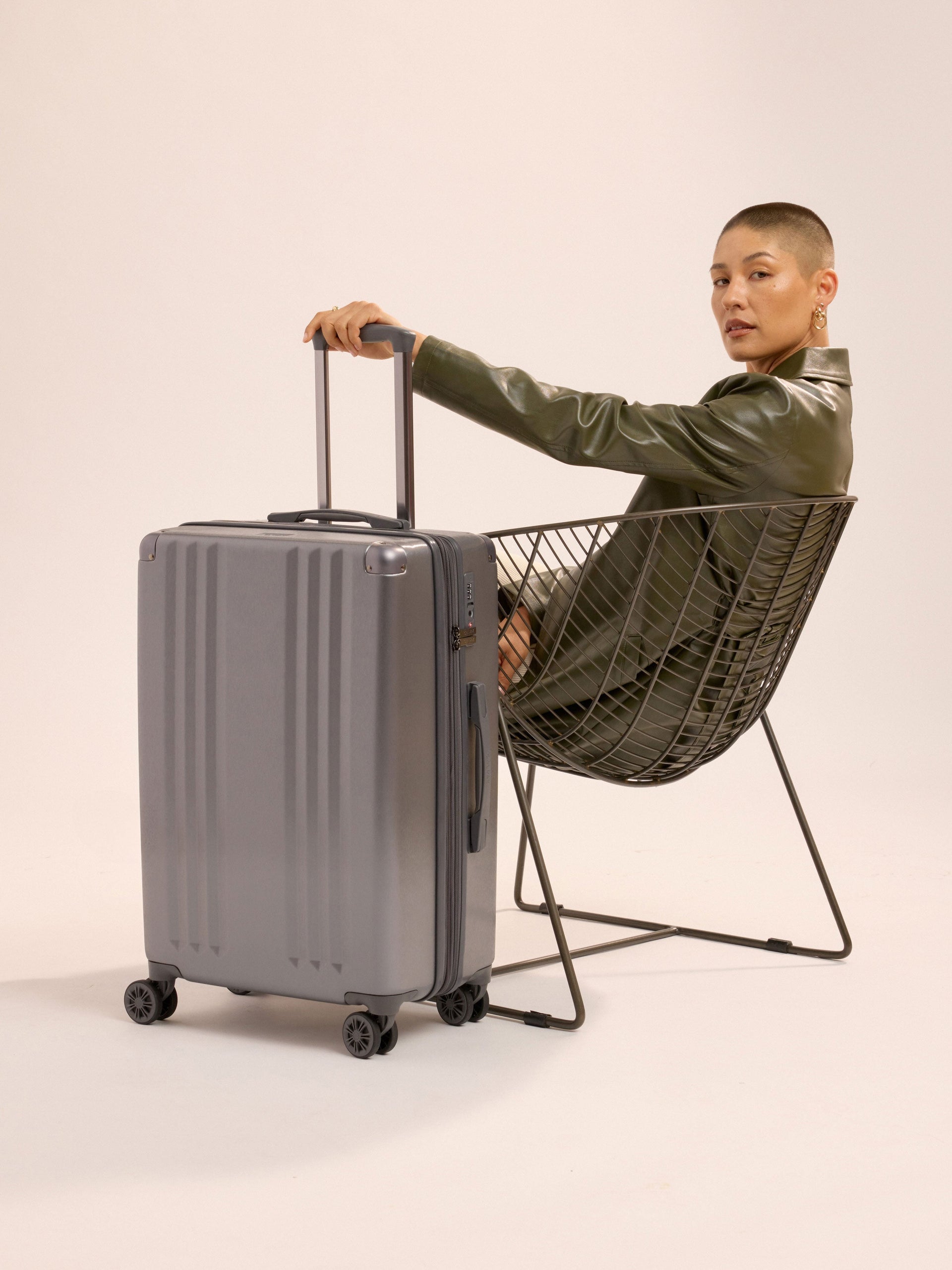 Model sitting besides CALPAK Ambeur Medium Luggage while holding telescopic trolley handle in metallic gunmetal gray; LAM1024-GUNMETAL