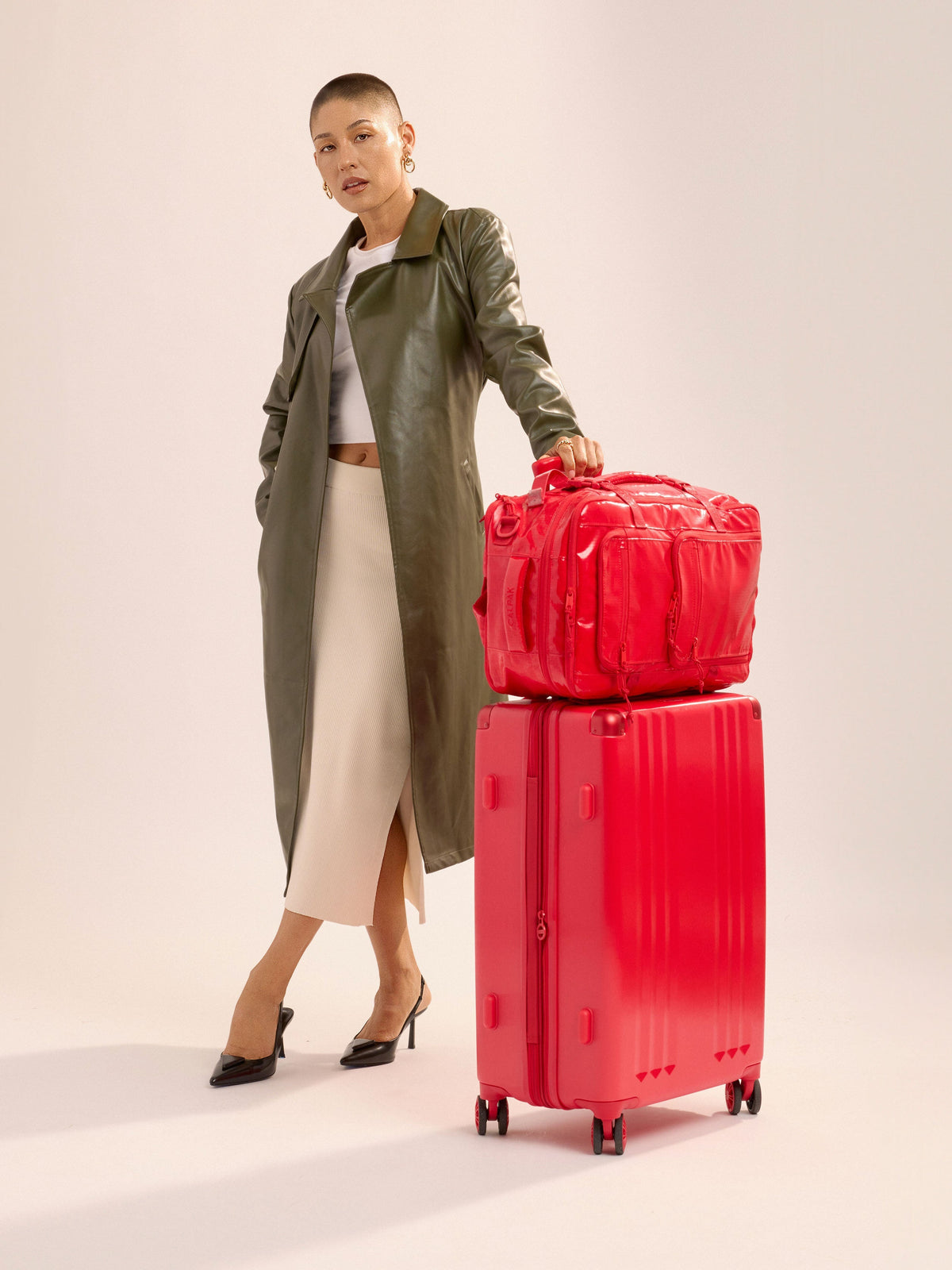 Model standing besides CALPAK Ambeur Medium Luggage with CALPAK Terra 26L Laptop Duffel Backpack attached by telescopic trolley handle in metallic red; LAM1024-METALLIC-RED