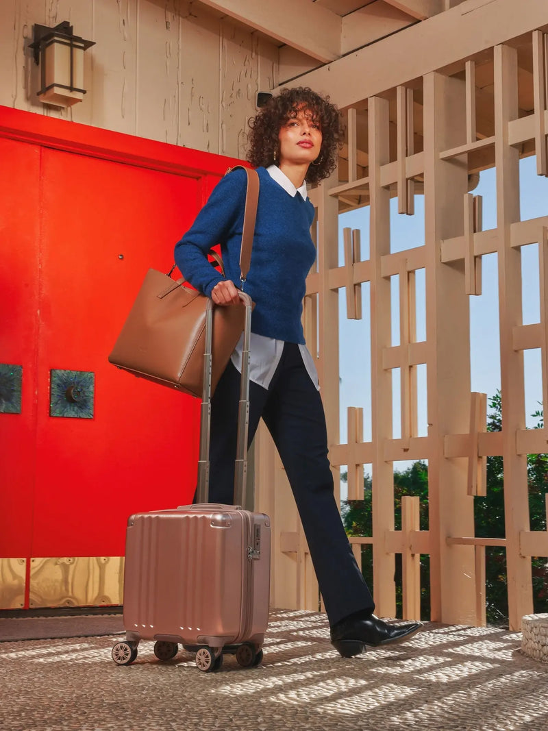 The CALPAK Ambuer Mini Carry-On suitcase in rose gold used by a model pulling it alongside her in an outdoor setting