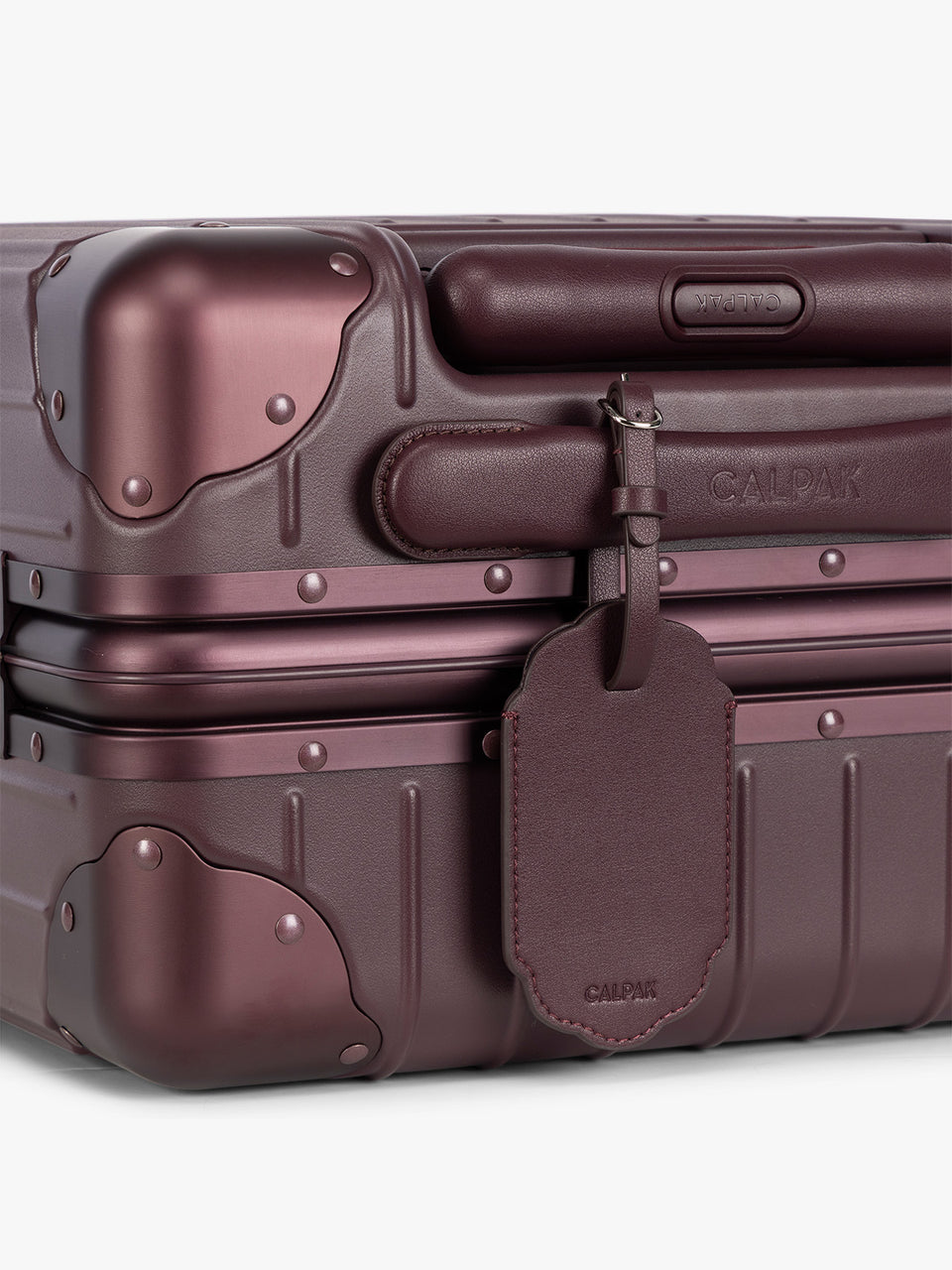 A close-up of a deep red hard-shell suitcase with protective corner bumpers, a deep red CALPAK Vault luggage tag with a scalloped border and a matching black strap, shown against a plain white background; ATV2601-DARK-CHERRY