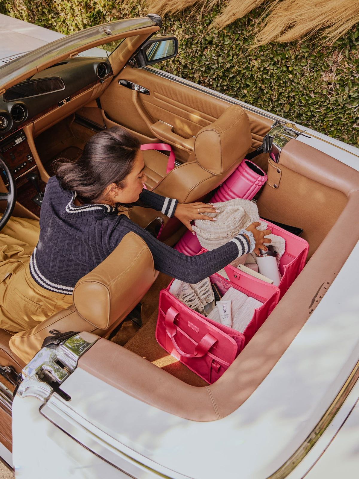 Model sitting in convertible car reaching towards the backseat for blanket in her CALPAK Car Organizer in dragonfruit; ACO2001-DRAGONFRUIT