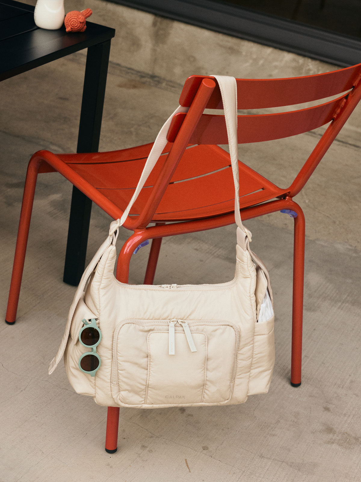 CALPAK Crossbody Diaper Bag in off-white Oatmeal hung on a red chair at a cafe; CDB2501-OATMEAL