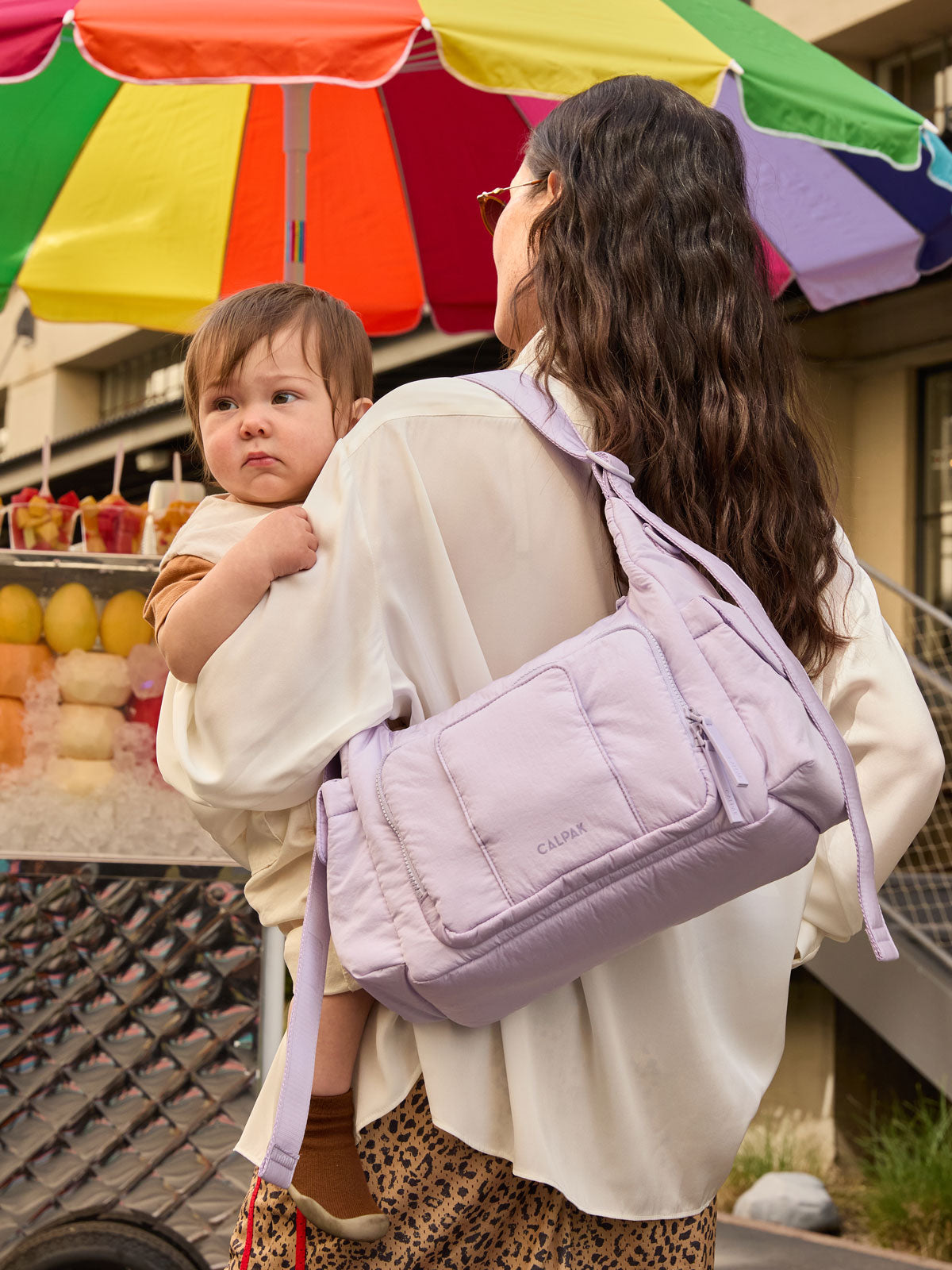 Model holding a baby and wearing the CALPAK Crossbody Diaper Bag in light purple Orchid; CDB2501-ORCHID