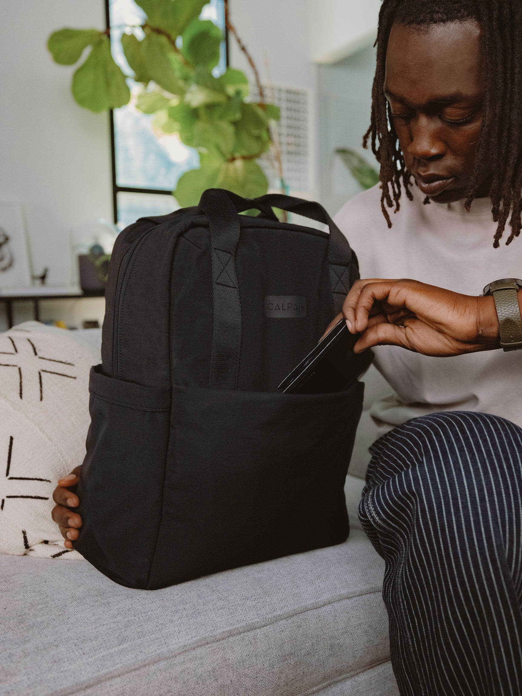 Model placing cell phone in front pocket of CALPAK Connect Laptop Backpack in black; BCN2401-BLACK