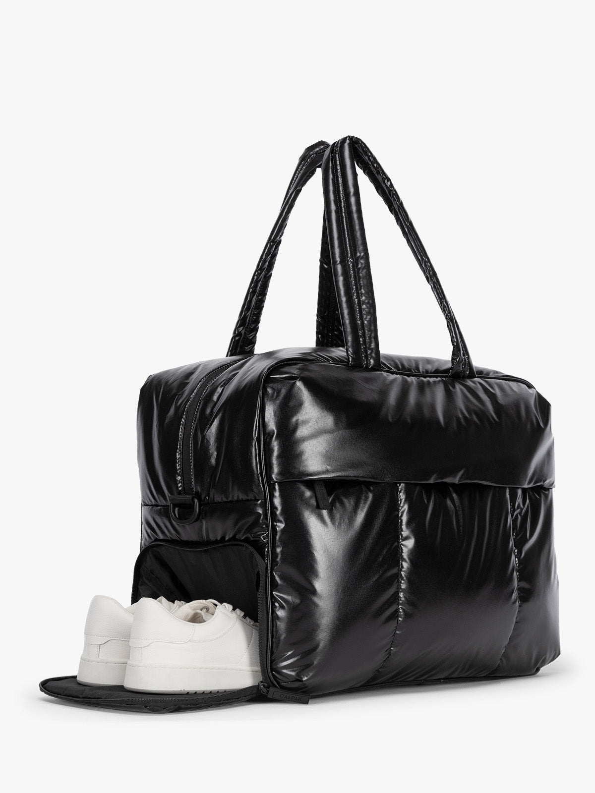 A shiny black puffer CALPAK Luka Large Duffel Bag with dual top handles, shown on a clean white background with a pair of white sneakers tucked inside the open lower front pocket; DLL2201-GLAZED-BLACK