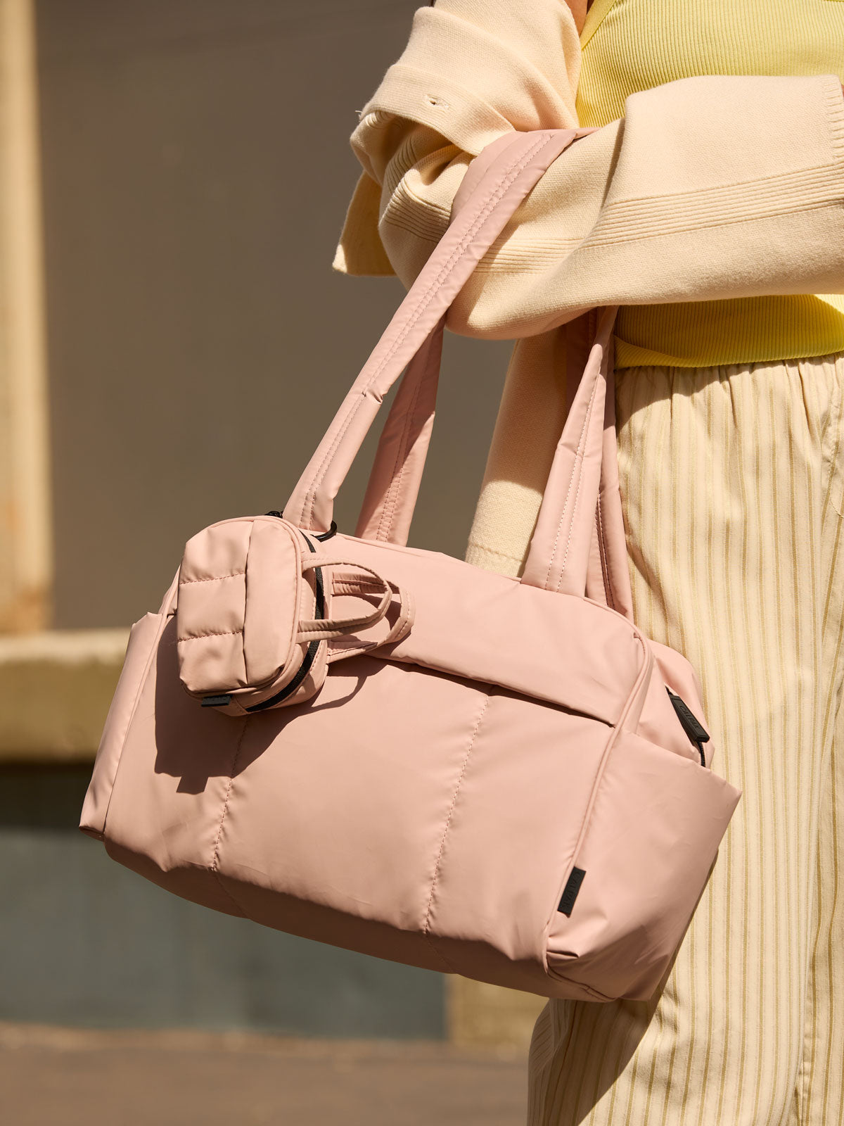 CALPAK Mini Duffel in pink Rose Quartz with puffy exterior, interior and exterior pockets, and padded shoulder straps being worn by a model; DLM2501-ROSE-QUARTZ
