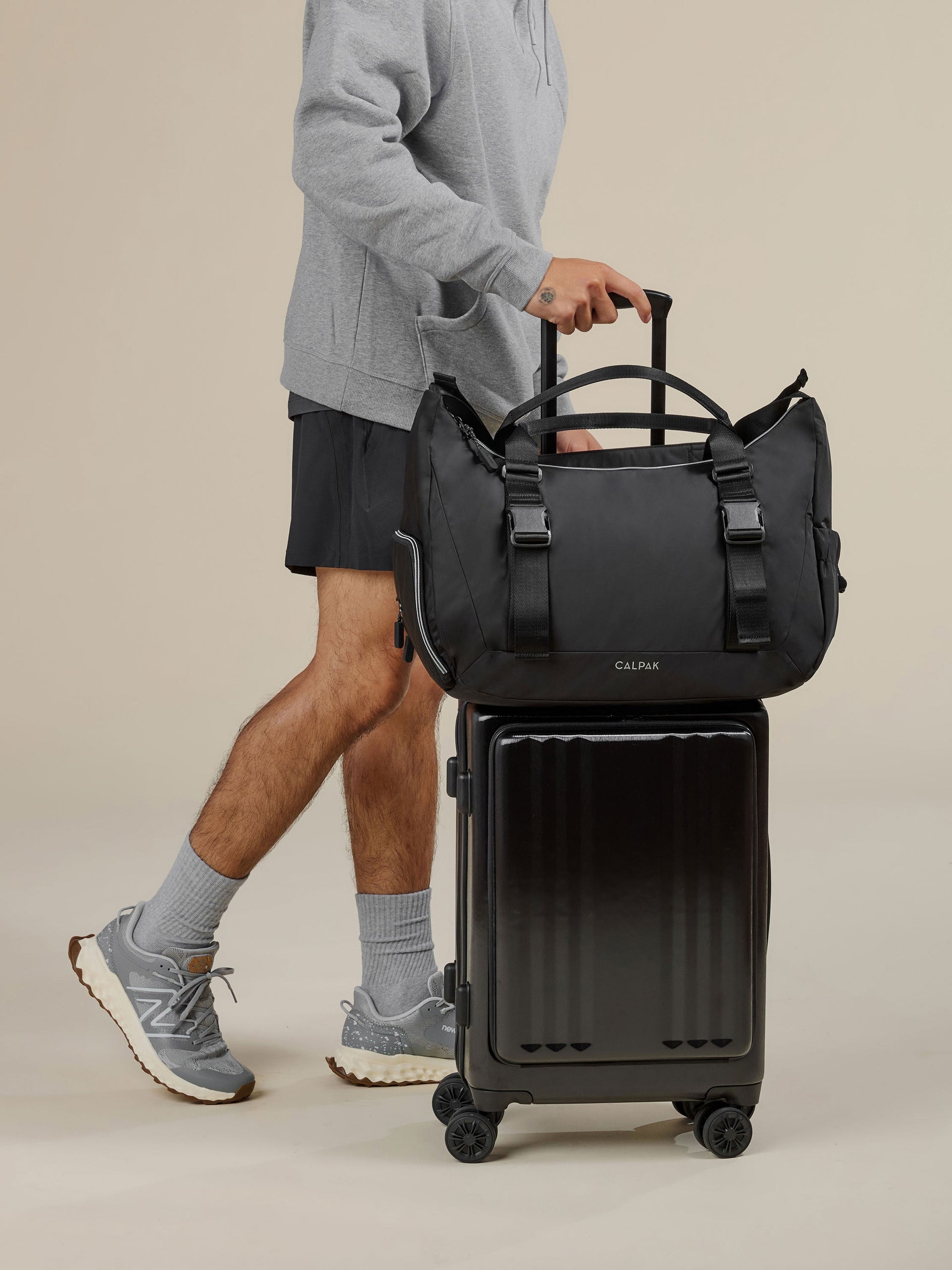 Model rolling CALPAK Ambeur Front Pocket Carry-On Luggage with CALPAK Fit Gym Bag secured by luggage trolley sleeve in black; TBF2401-BLACK, BTBF2401-BLACK