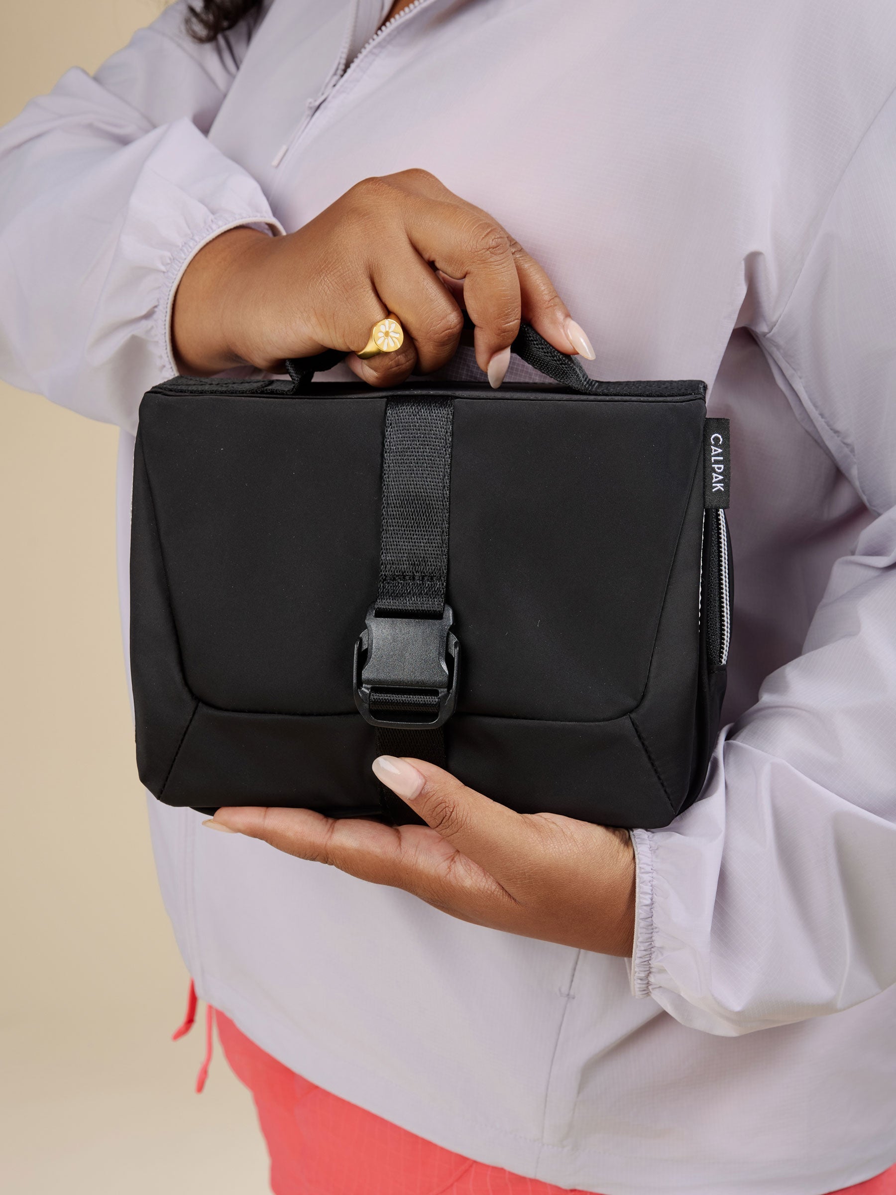 Model holding CALPAK Fit hanging gym toiletry bag with front buckle closure and top carrying handle in black; TFH2401-BLACK, TFH2401-BLACK-GRIN, BTBF2401-BLACK