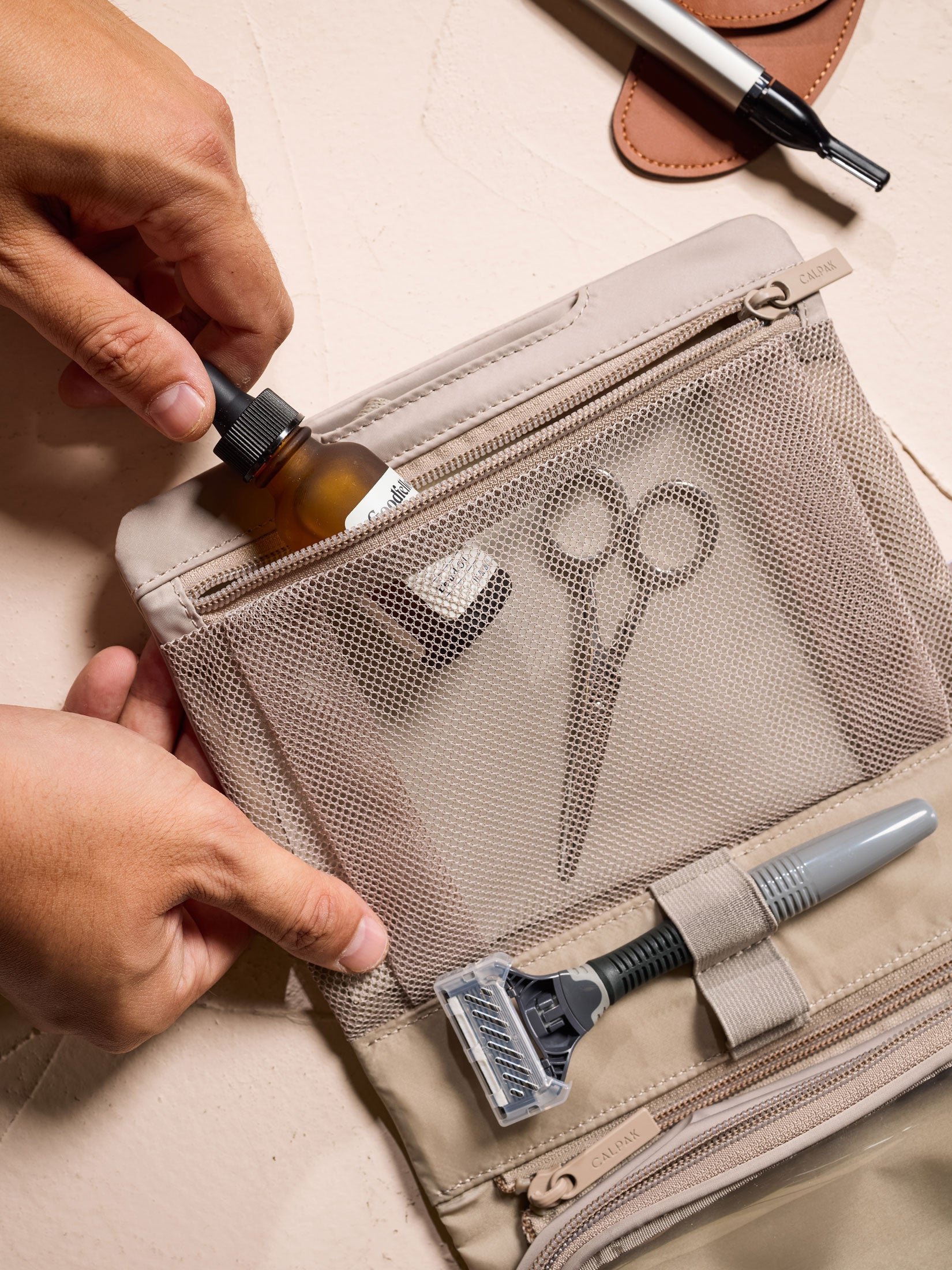 Model placing small toiletry item within interior mesh pocket of CALPAK Fit Hanging Dopp Kit in greige; TFH2401-GREIGE, TFH2401-GREIGE-GRIN, BTBF2401-GREIGE