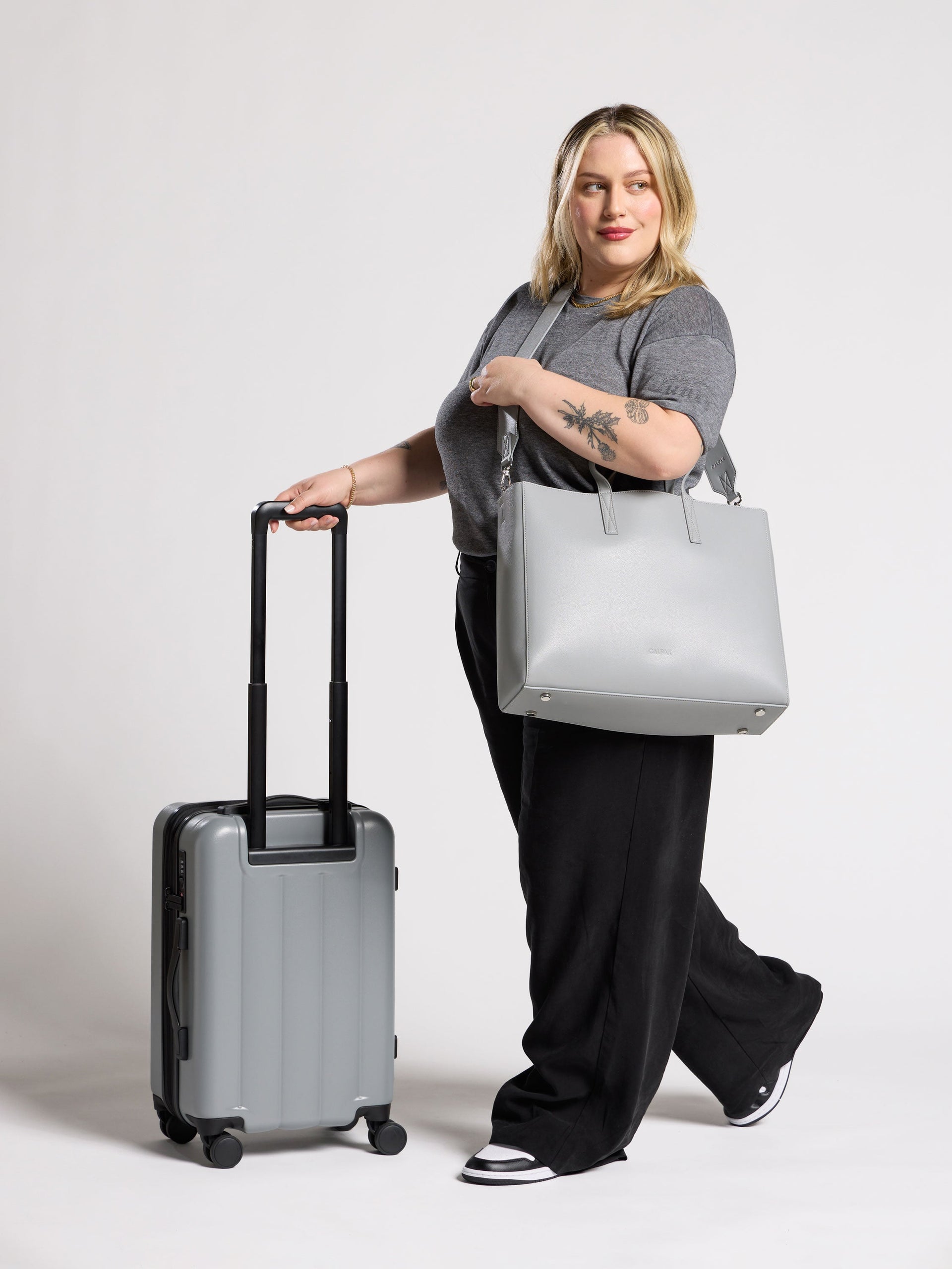 Model wearing CALPAK Haven Laptop Tote over shoulder while rolling Evry Carry-On Luggage in smoke; ATO2101-SMOKE