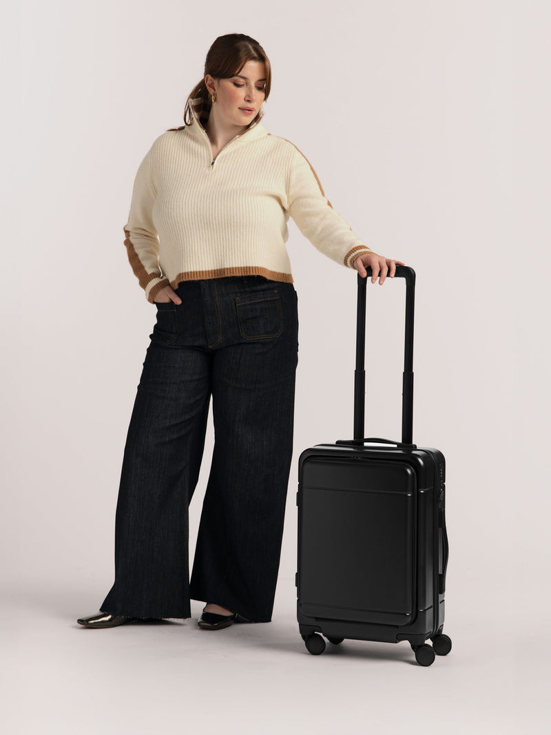 Model standing with Hue Front Pocket Carry-On Luggage in Black with handle extended and 360 spinner wheels