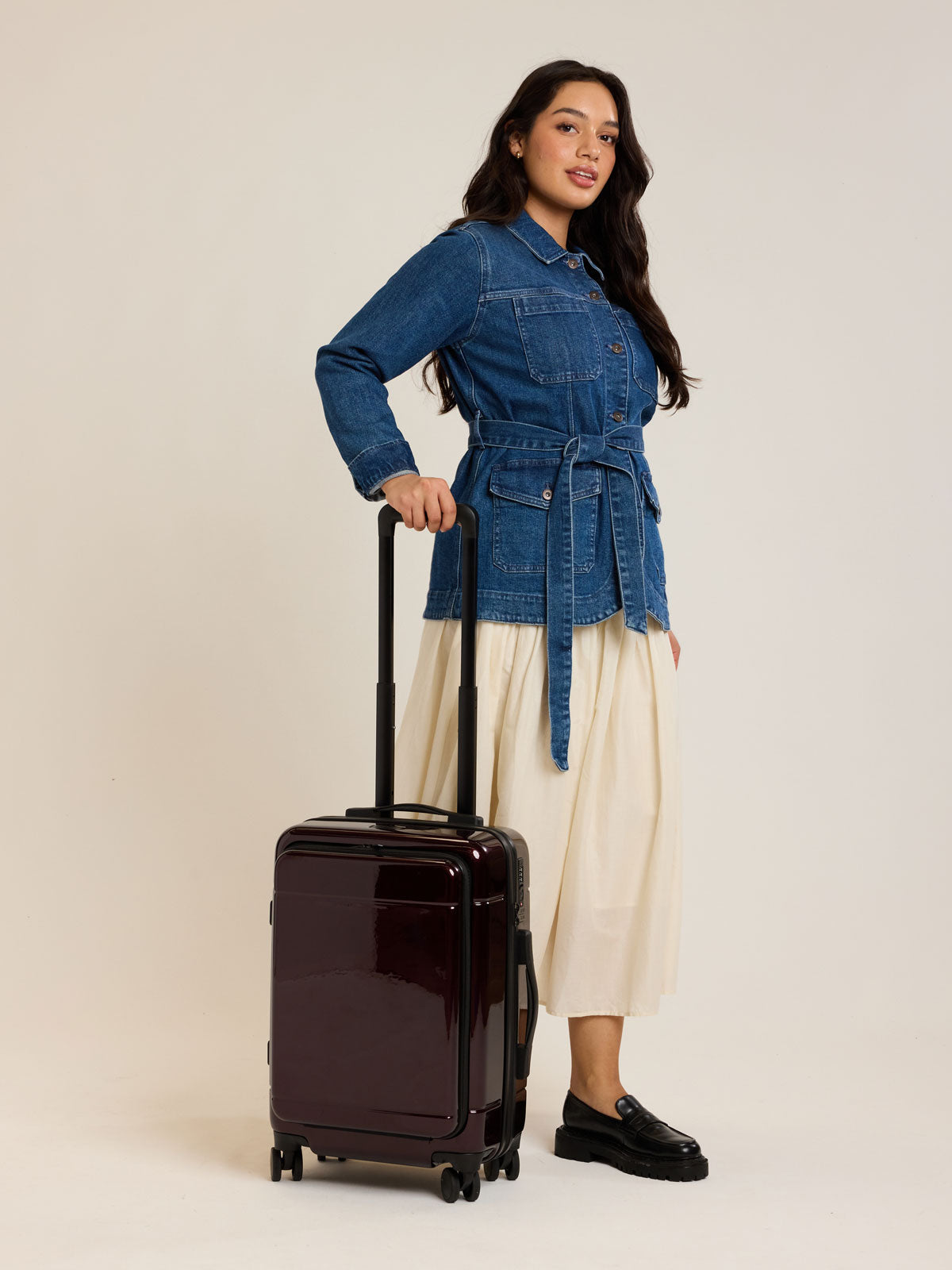 Model holding CALPAK Hue Front Pocket Carry On Luggage in maroon Glazed Dark Cherry ; LHU1020-GLAZED-DARK-CHERRY