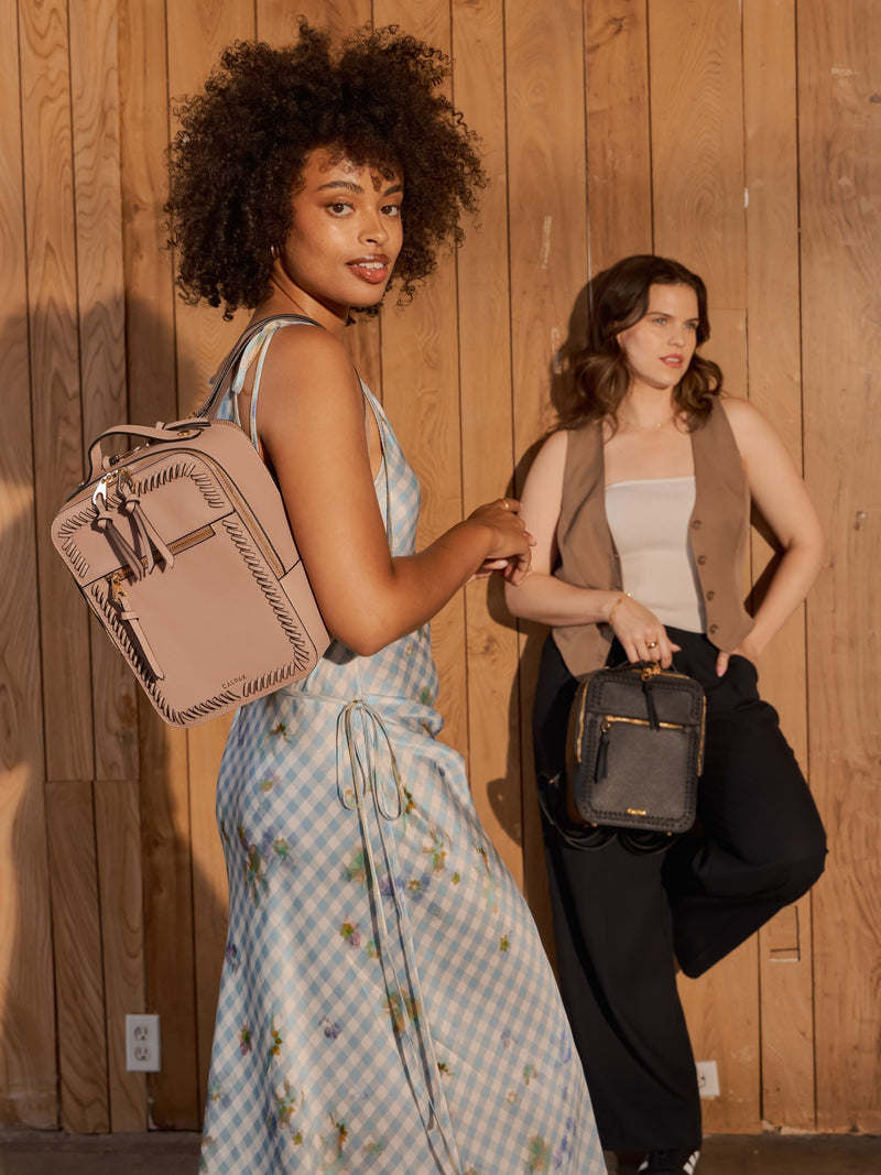 A woman carries a Kaya mauve mini backpack over one shoulder while another woman carries the Kaya mini backpack in black by the hand