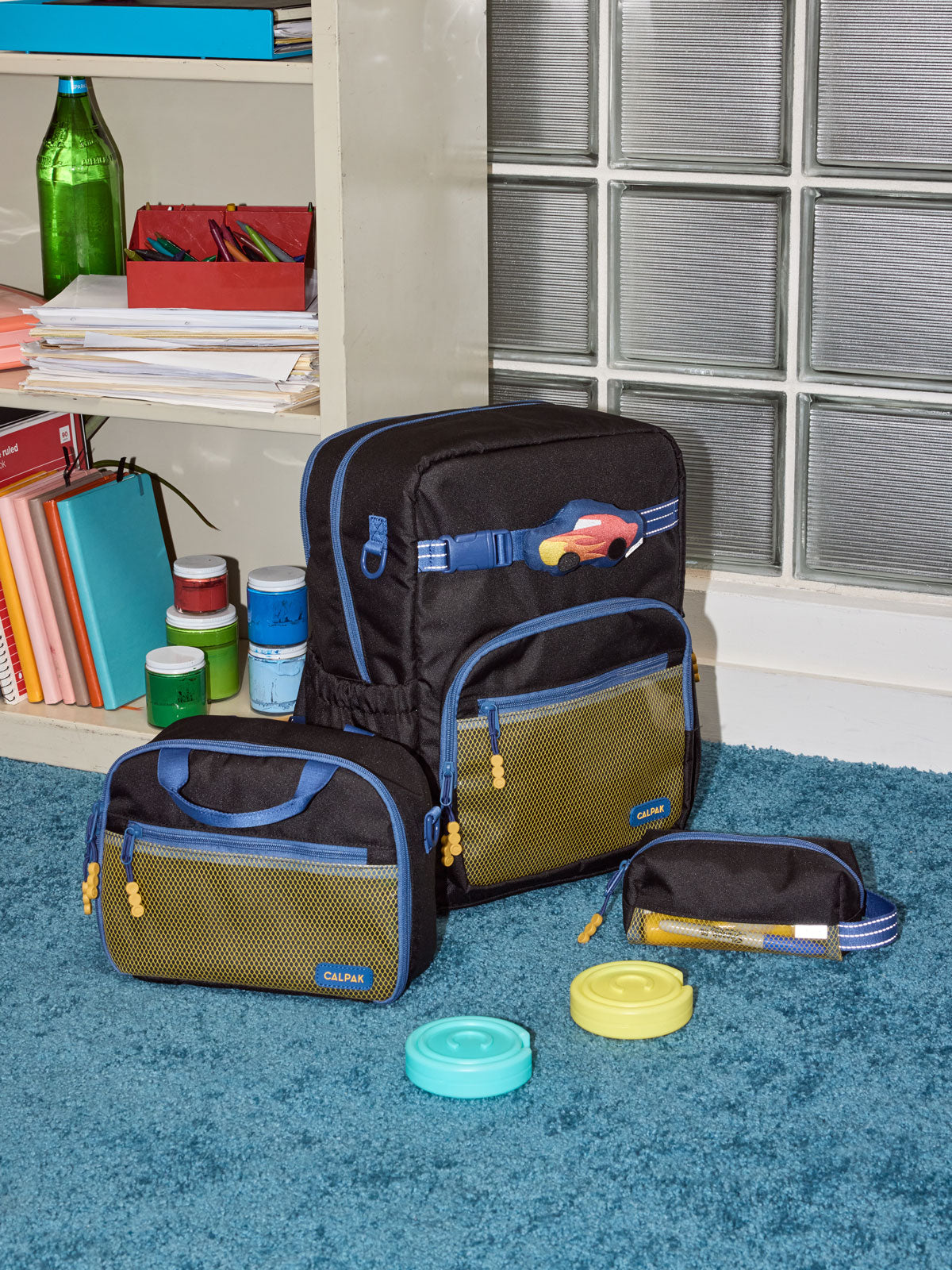 CALPAK Kids Collection Large Kids Backpack Bundle in Black that includes Kids Lunch Bag, Kids Pencil Case, red Car Pluffy Bag Charm, and Set of 2 Ice Packs in a kids room; BCKL5000-BLACK