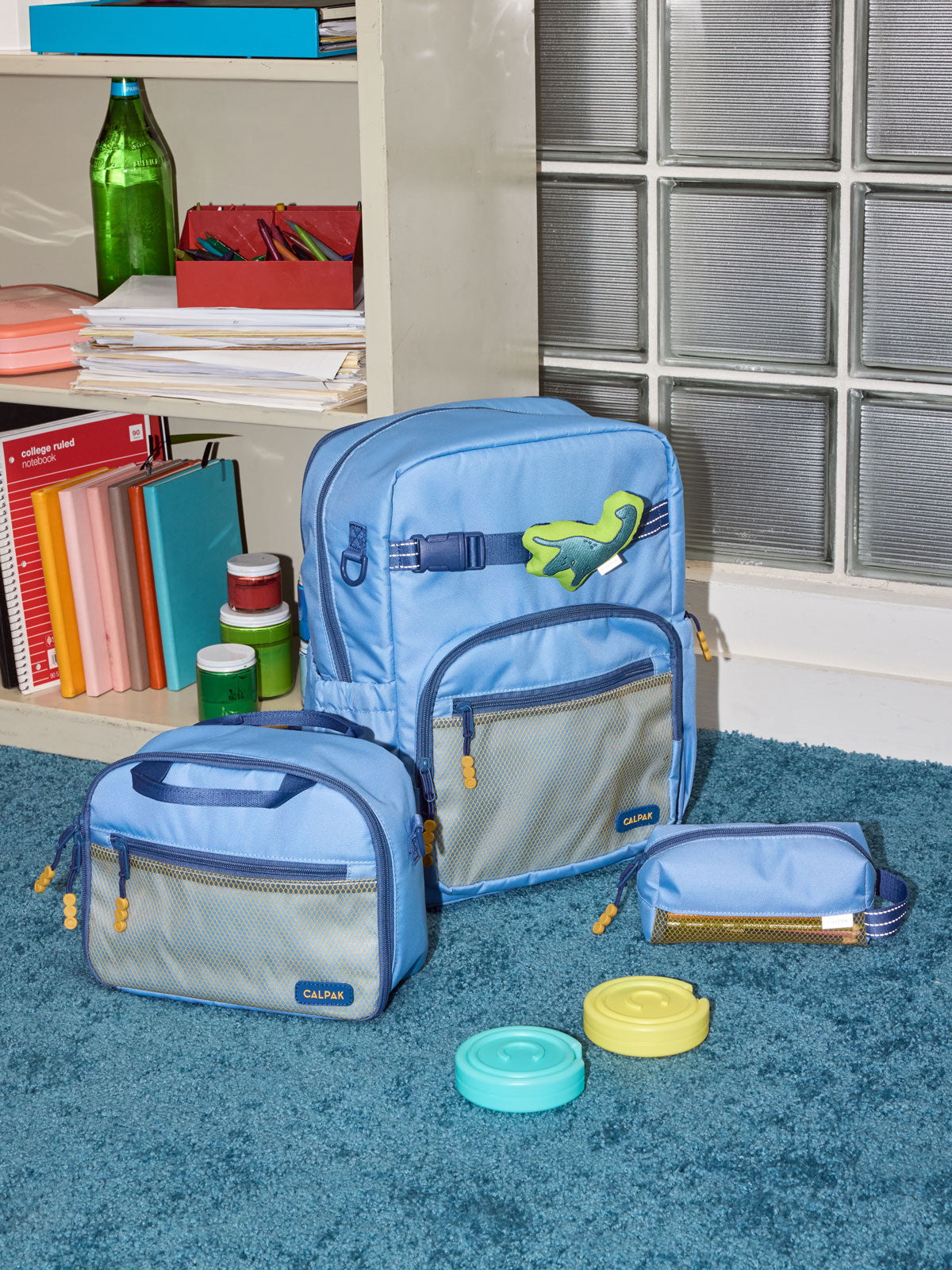 CALPAK Kids Collection Large Kids Backpack Bundle in Blue that includes Kids Lunch Bag, Kids Pencil Case, Rainbow Pluffy Bag Charm, and Set of 2 Ice Packs in a kid's room; BCKL5000-BLUE