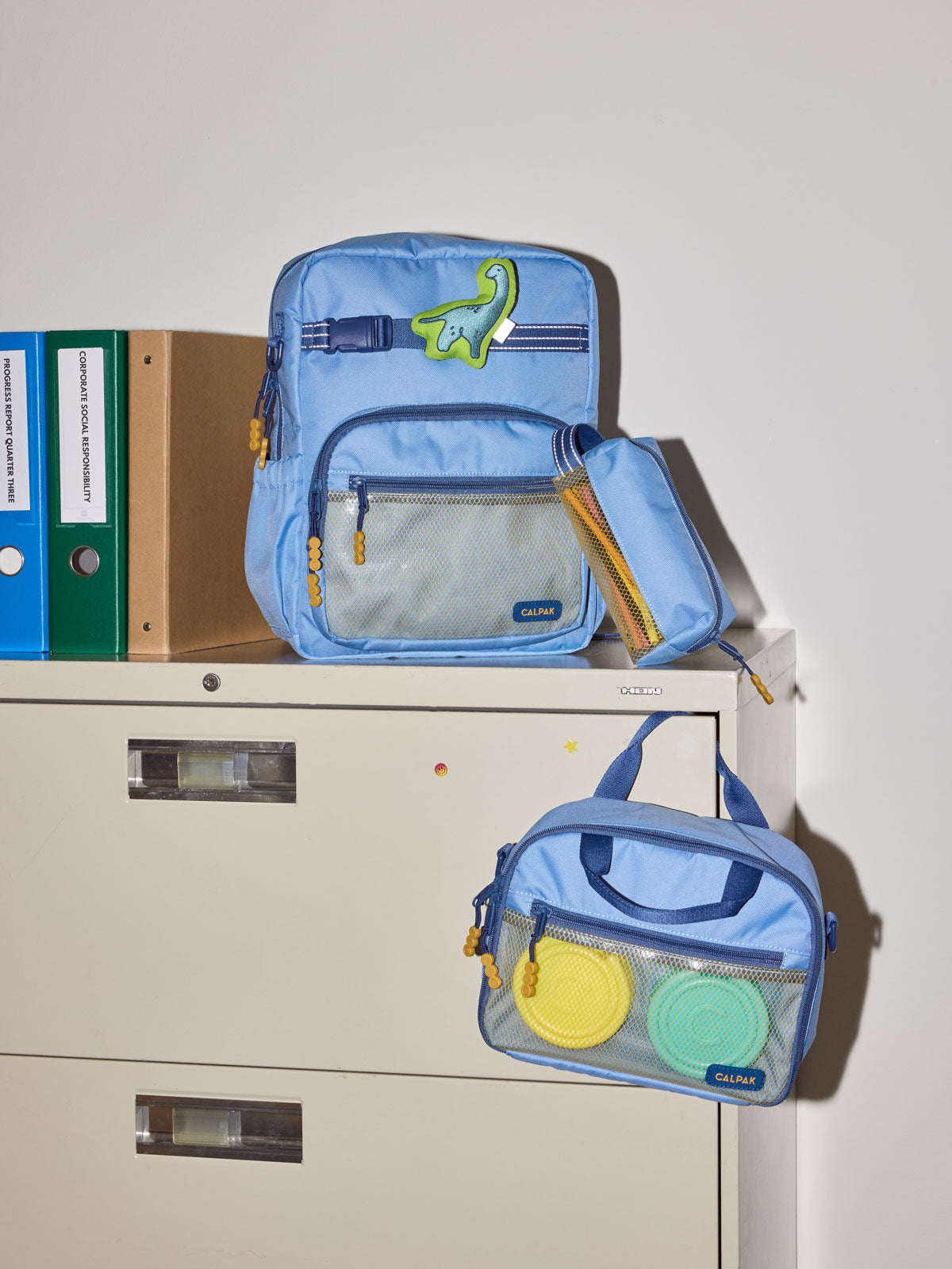 CALPAK Kids Collection Medium Kids Backpack Bundle in Blue that includes Kids Lunch Bag, Kids Pencil Case, blue and green Dinosaur Pluffy Bag Charm, and Set of 2 Ice Packs in a kid's room; BCKM5000-BLUE