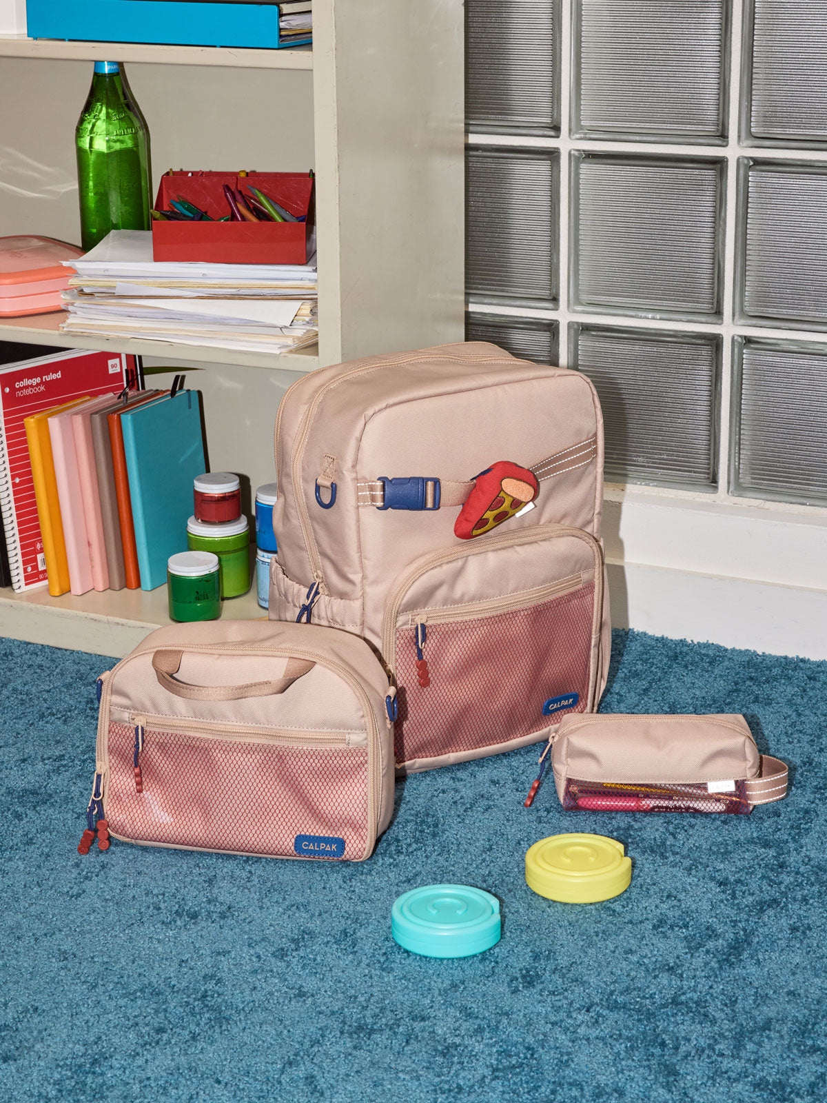 CALPAK Kids Collection Large Kids Backpack Bundle in brown/tan Khaki that includes Kids Lunch Bag, Kids Pencil Case, red and yellow Pizza Pluffy Bag Charm, and Set of 2 Ice Packs in a kid's room; BCKL5000-KHAKI