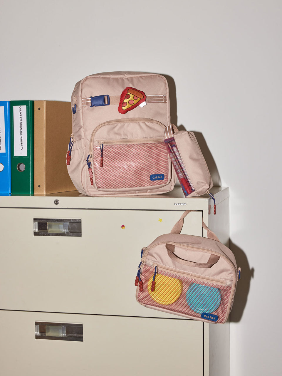 CALPAK Kids Medium Backpack in brown and tan Khaki for ages 5-8 is built for your little one’s big days with dark brown and tan zippers and red mesh front pocket, multiple interior pockets, and padded comfort straps on a beige cabinet; CKBM2501-KHAKI