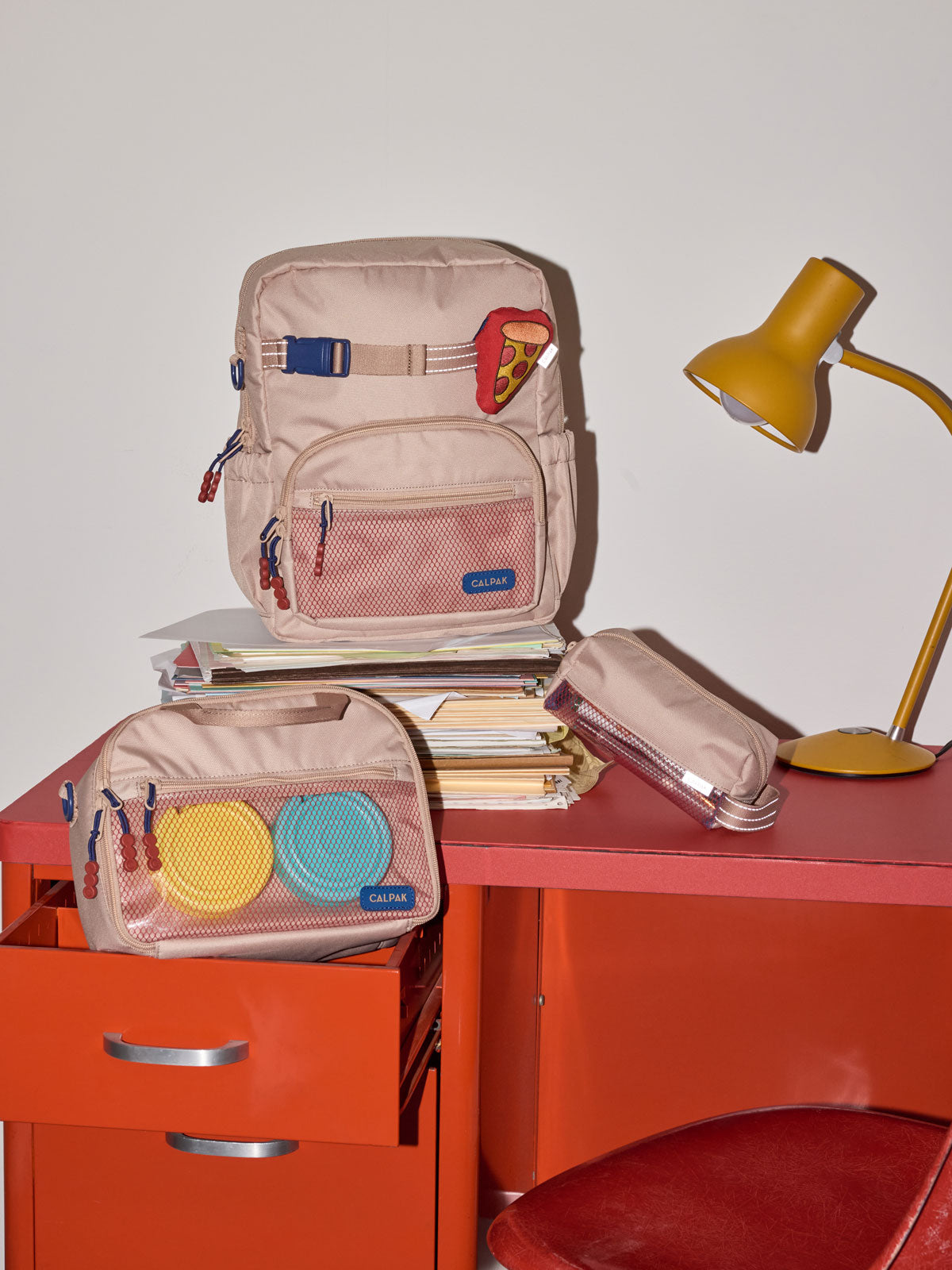 CALPAK Kids Collection Small Kids Backpack Bundle in brown/tan Khaki that includes Kids Lunch Bag, Kids Pencil Case, red and yellow PIzza Pluffy Bag Charm, and Set of 2 Ice Packs in a kid's room; BCKS5000-KHAKI