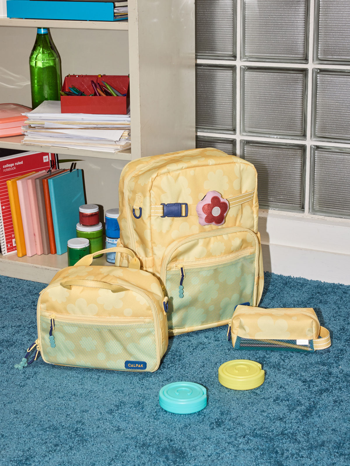 CALPAK Kids Collection Large Kids Backpack Bundle in yellow flower print Yellow Bloom that includes Kids Lunch Bag, Kids Pencil Case, red Car Pluffy Bag Charm, and Set of 2 Ice Packs in a kid's room; BCKL5000-YELLOW-BLOOM