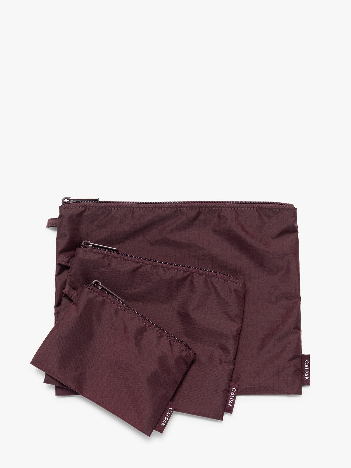 Three burgundy Dark Cherry CALPAK Compakt Zippered pouches of different sizes, arranged in a fan-like layout on a clean white background; KZB2001-DARK-CHERRY