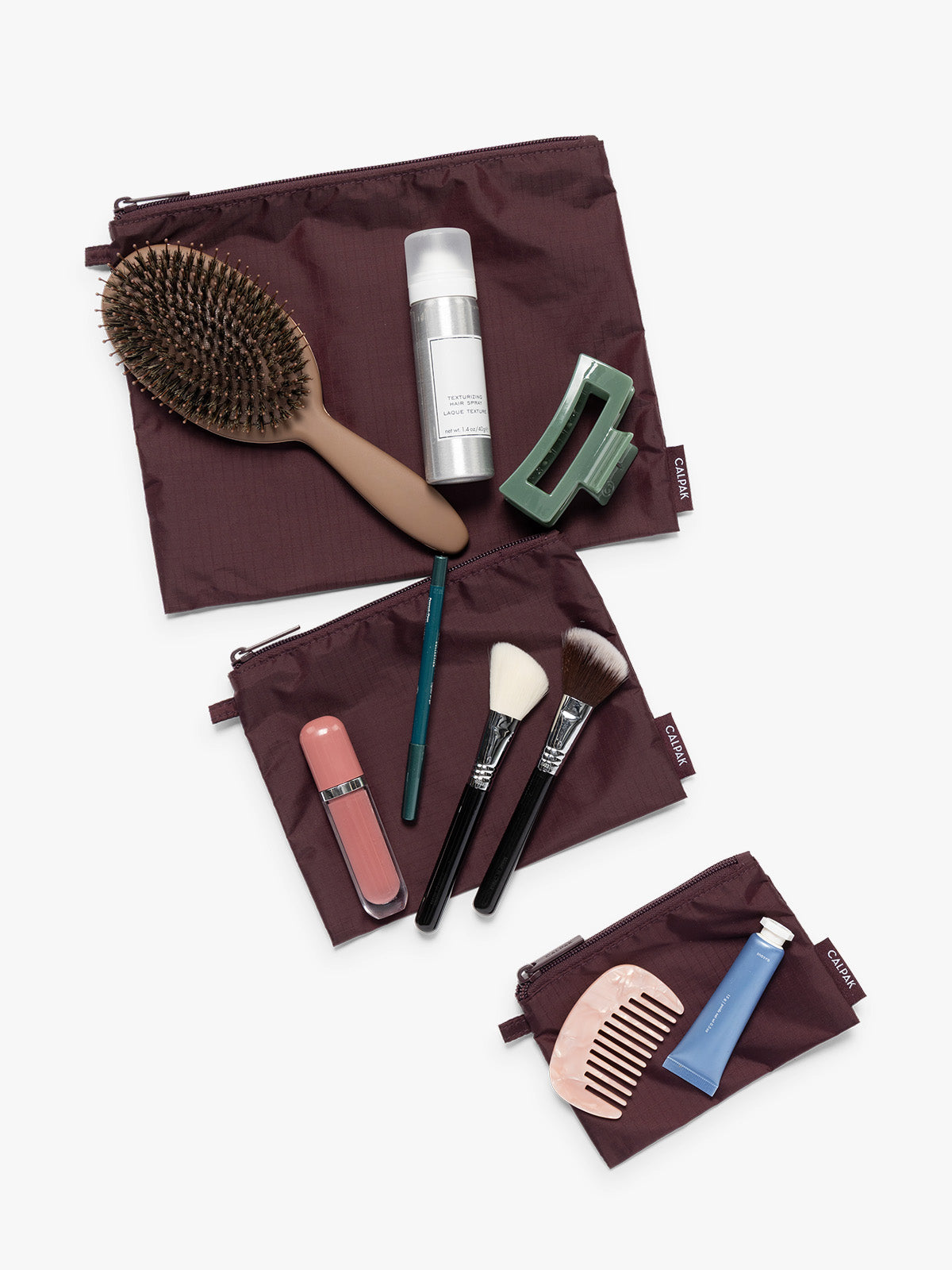 Items on top of the three burgundy Dark Cherry CALPAK Compakt Zippered pouches of different sizes, with items on top showing what fits inside, on a clean white background; KZB2001-DARK-CHERRY