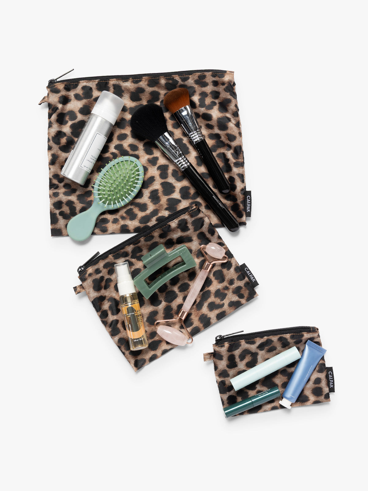 Items on top of the three tan and black animal print Leopard CALPAK Compakt Zippered pouches of different sizes, with items on top showing what fits inside, on a clean white background; KZB2001-LEOPARD