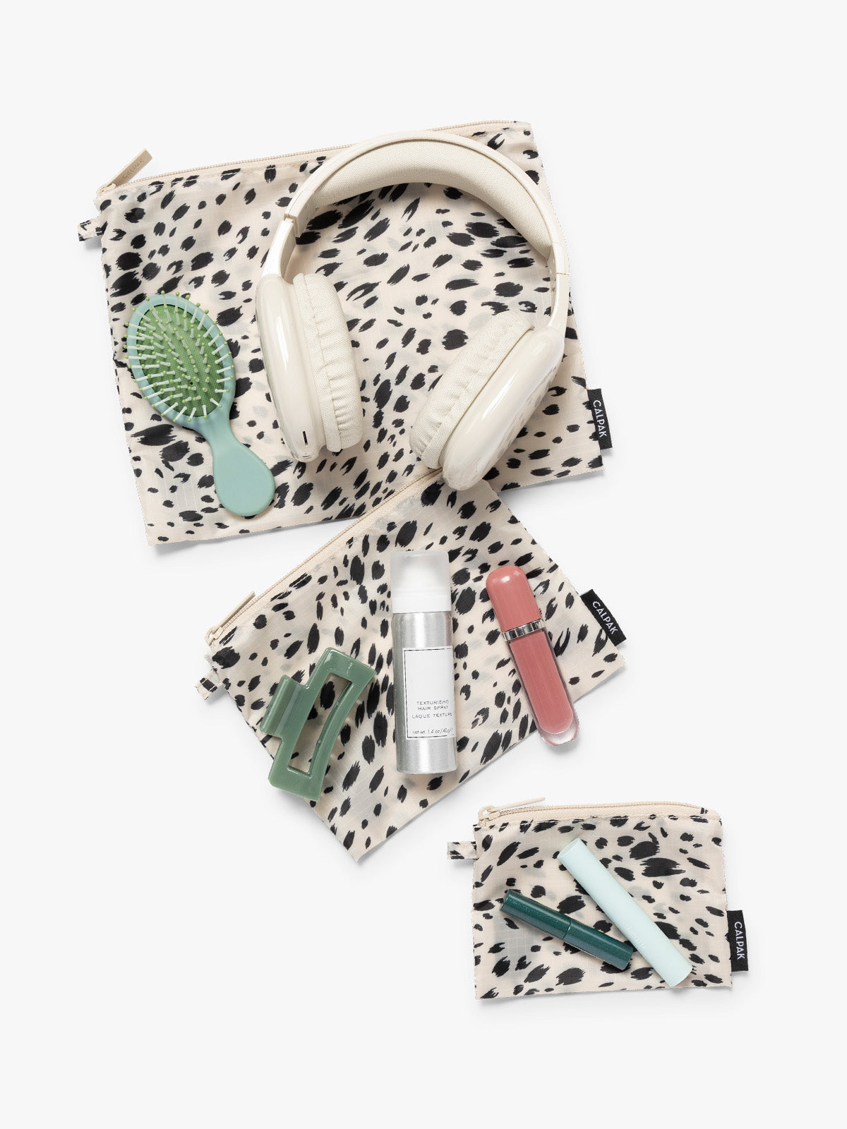 Items on top of the three white and black animal print Snow Leopard CALPAK Compakt Zippered pouches of different sizes, with items on top showing what fits inside, on a clean white background; KZB2001-SNOW-LEOPARD