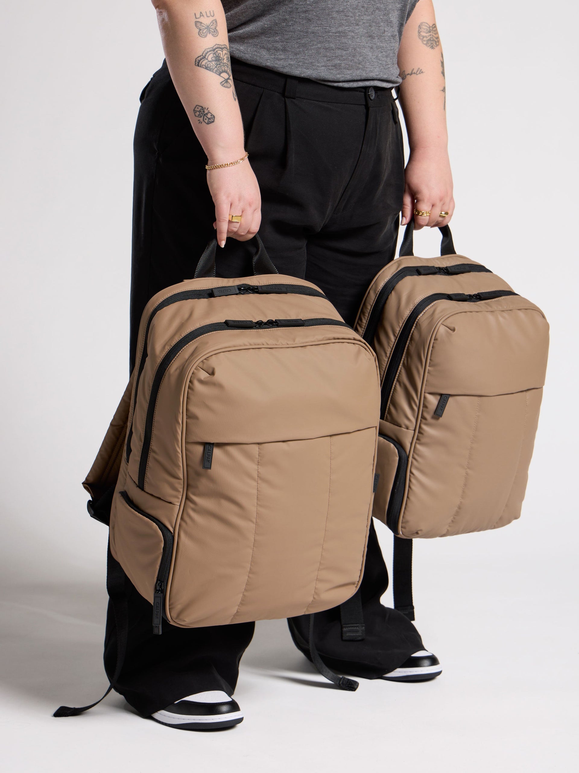 Model holding CALPAK 17 inch Laptop Backpack and CALPAK 15 inch Laptop Backpack comparing size difference in chocolate; BPX2301-CHOCOLATE