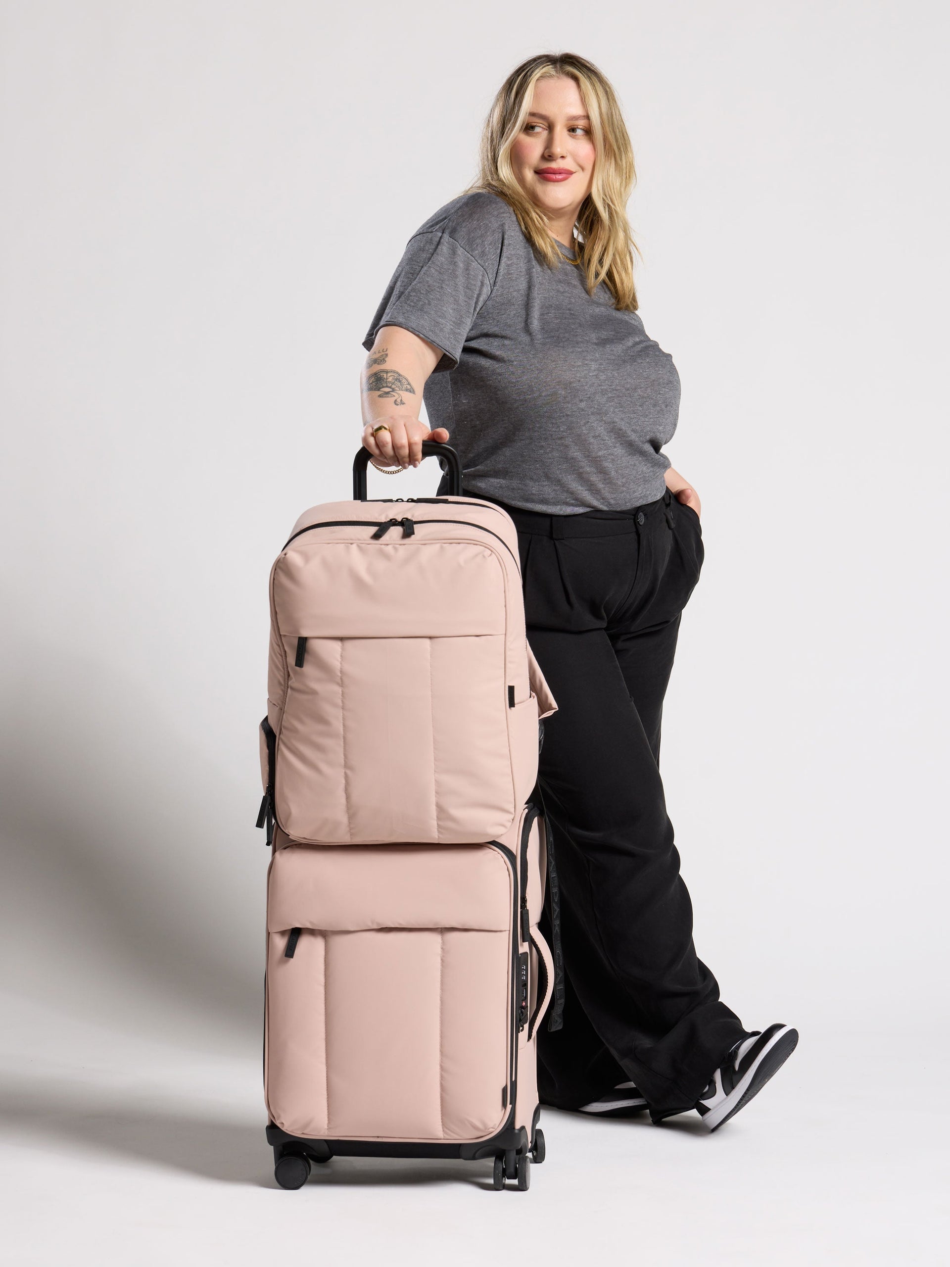 Model standing besides CALPAK 17 inch Laptop Backpack secured to Luka Soft-Sided Carry-On Luggage by trolley pass through in rose quartz; BPX2301-ROSE-QUARTZ