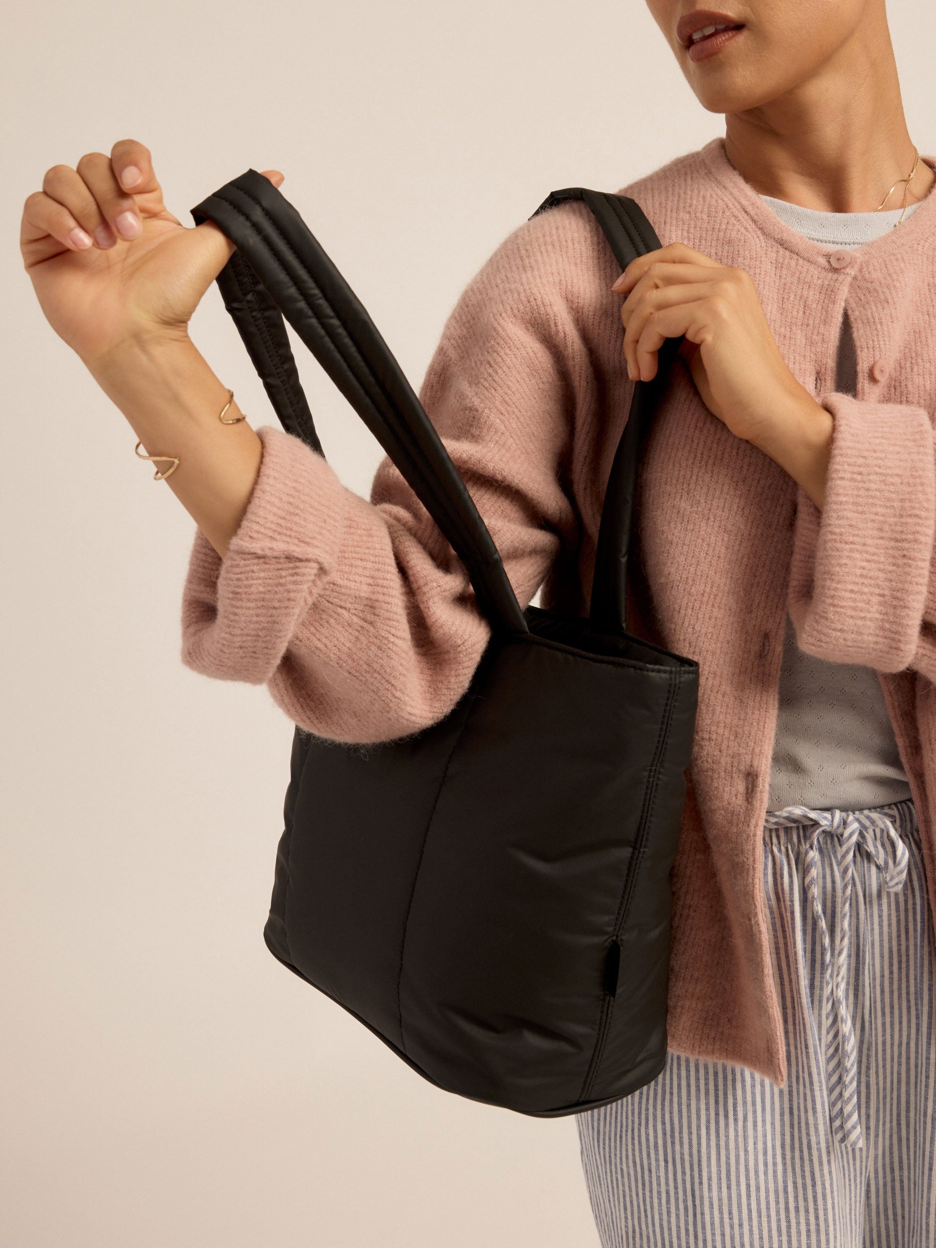 Model wearing black CALPAK Luka Mini Tote over shoulder with thumb holding durable padded shoulder strap; ALM2401-MATTE-BLACK