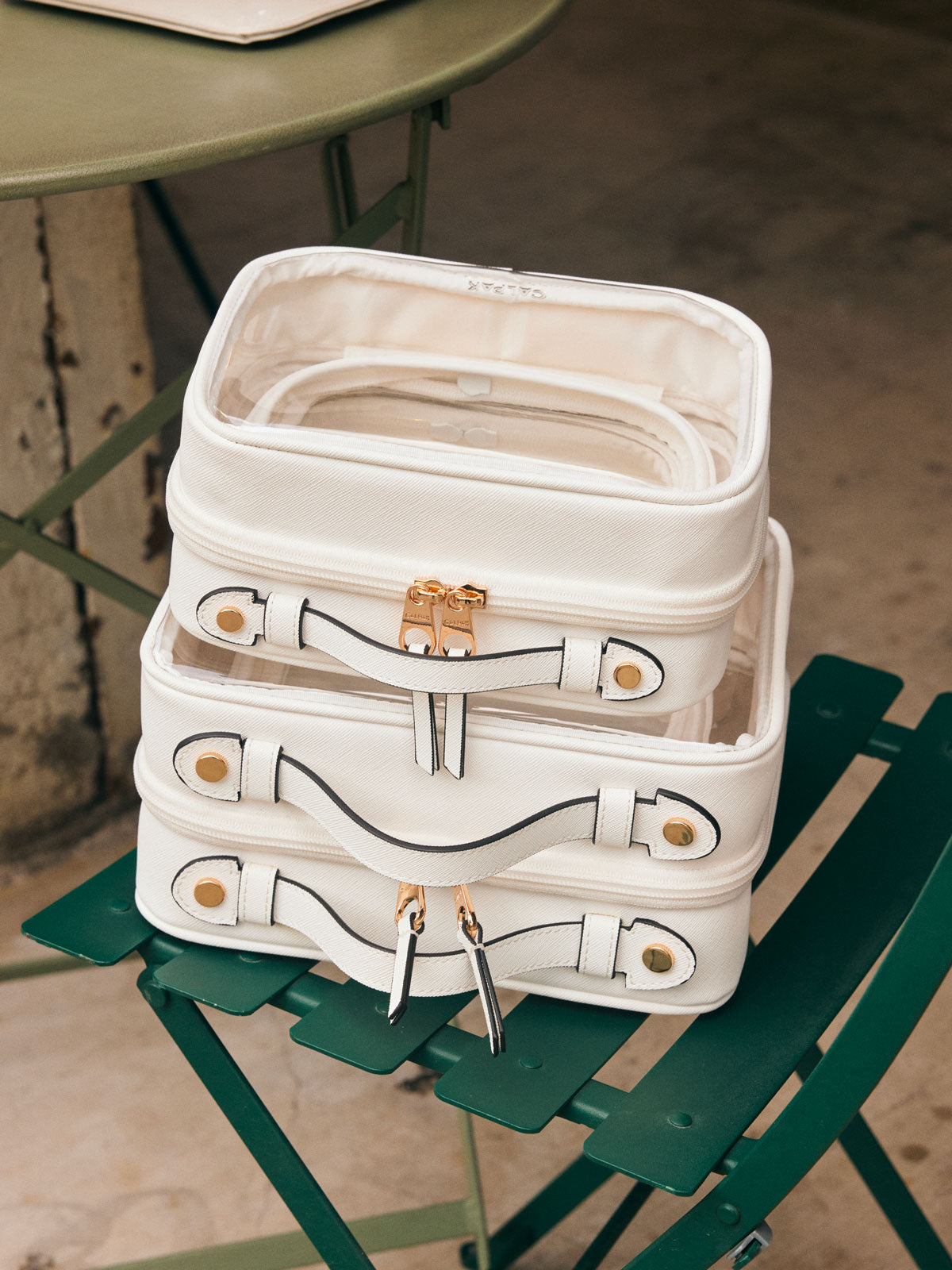 CALPAK Small, Medium, Large Clear Cosmetic Cases in White on a green chair, stacked on top of each other; CMM2201-WHITE