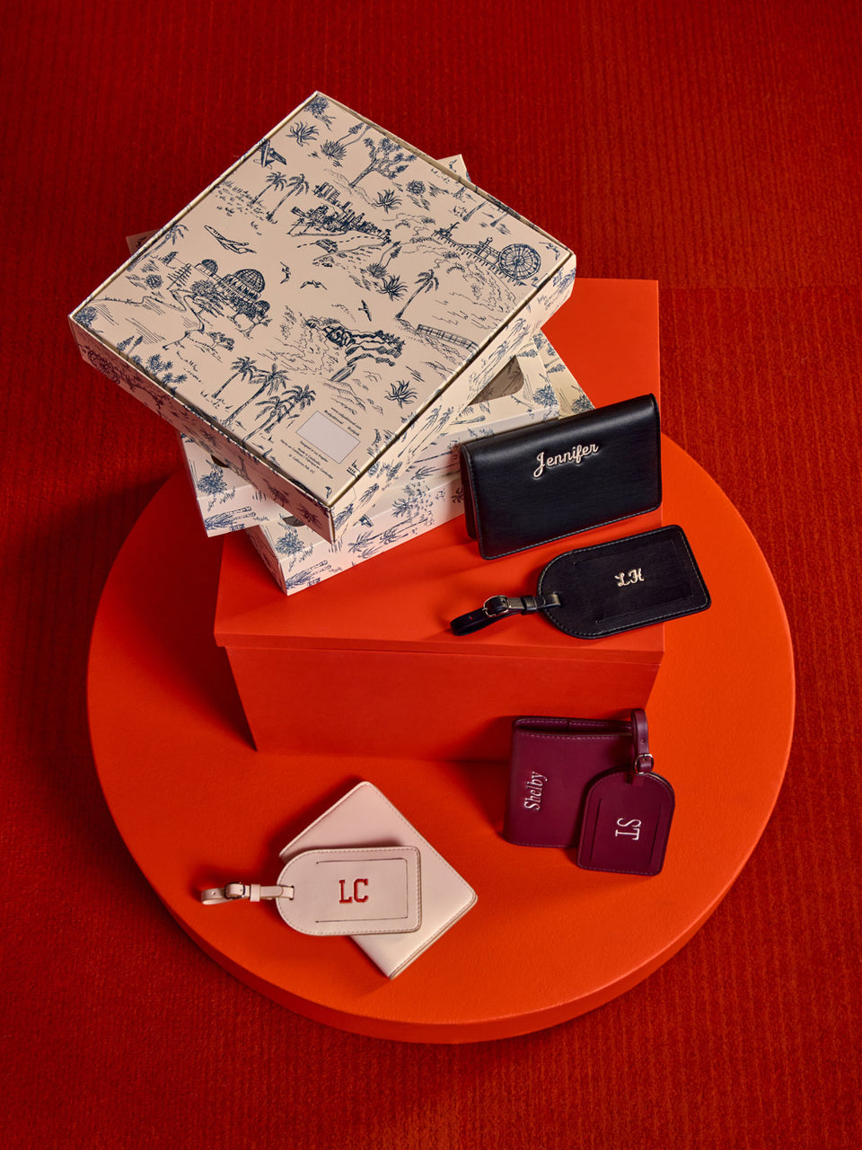 A styled flat-lay of CALPAK Luggage Tag and Passport Holder Set. Showing gift boxes arranged on a circular red platform against a textured red background. The set includes a cream toile-print box, black tags with gold monogram initials, and two burgundy tags with purple monogram initials; PWTS2601-BLACK