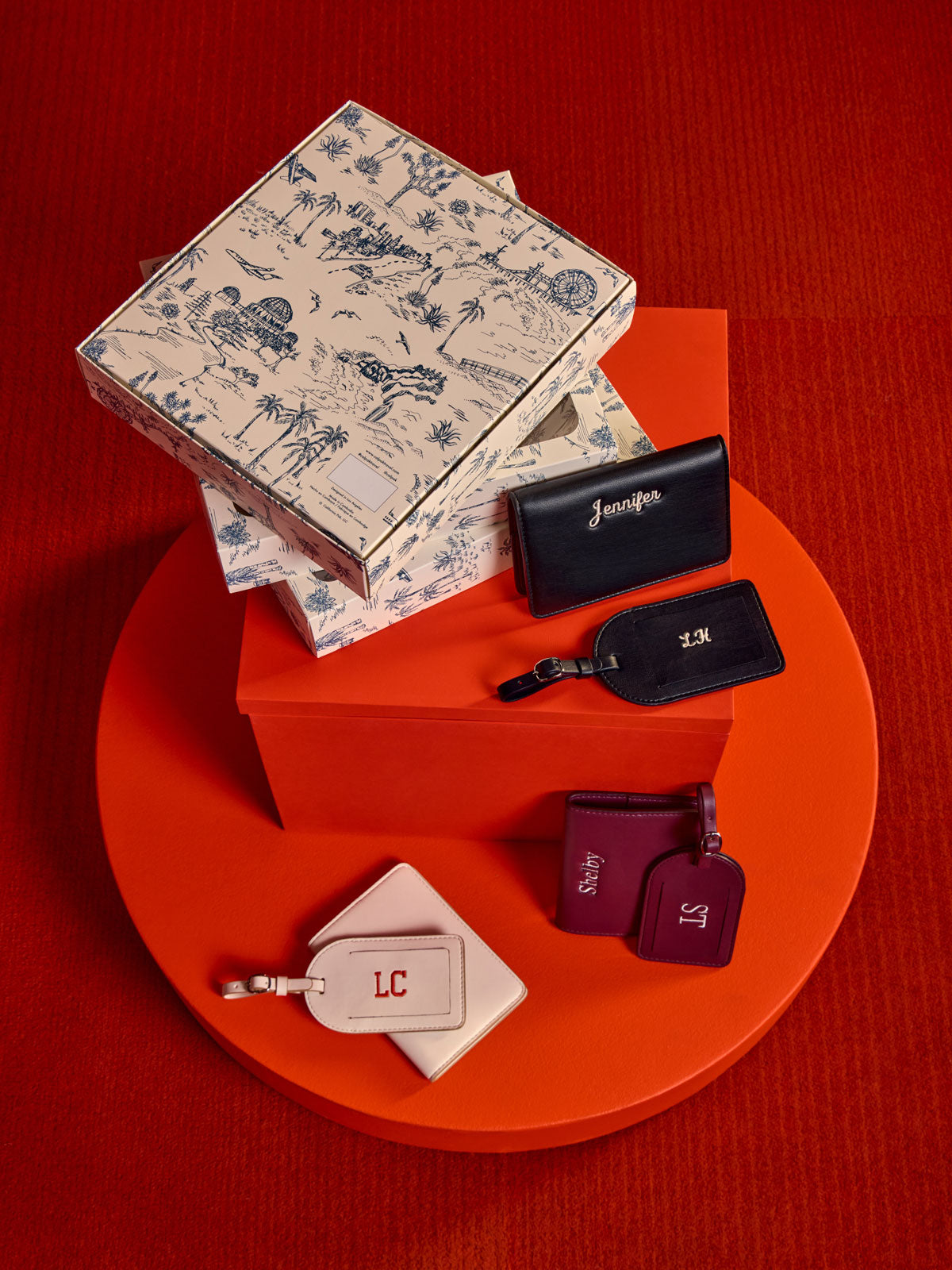 A styled flat-lay of CALPAK Luggage Tag and Passport Holder Set. Showing gift boxes arranged on a circular red platform against a textured red background. The set includes a cream toile-print box, black tags with gold monogram initials, and two burgundy tags with purple monogram initials;  PWTS2601-VANILLA
