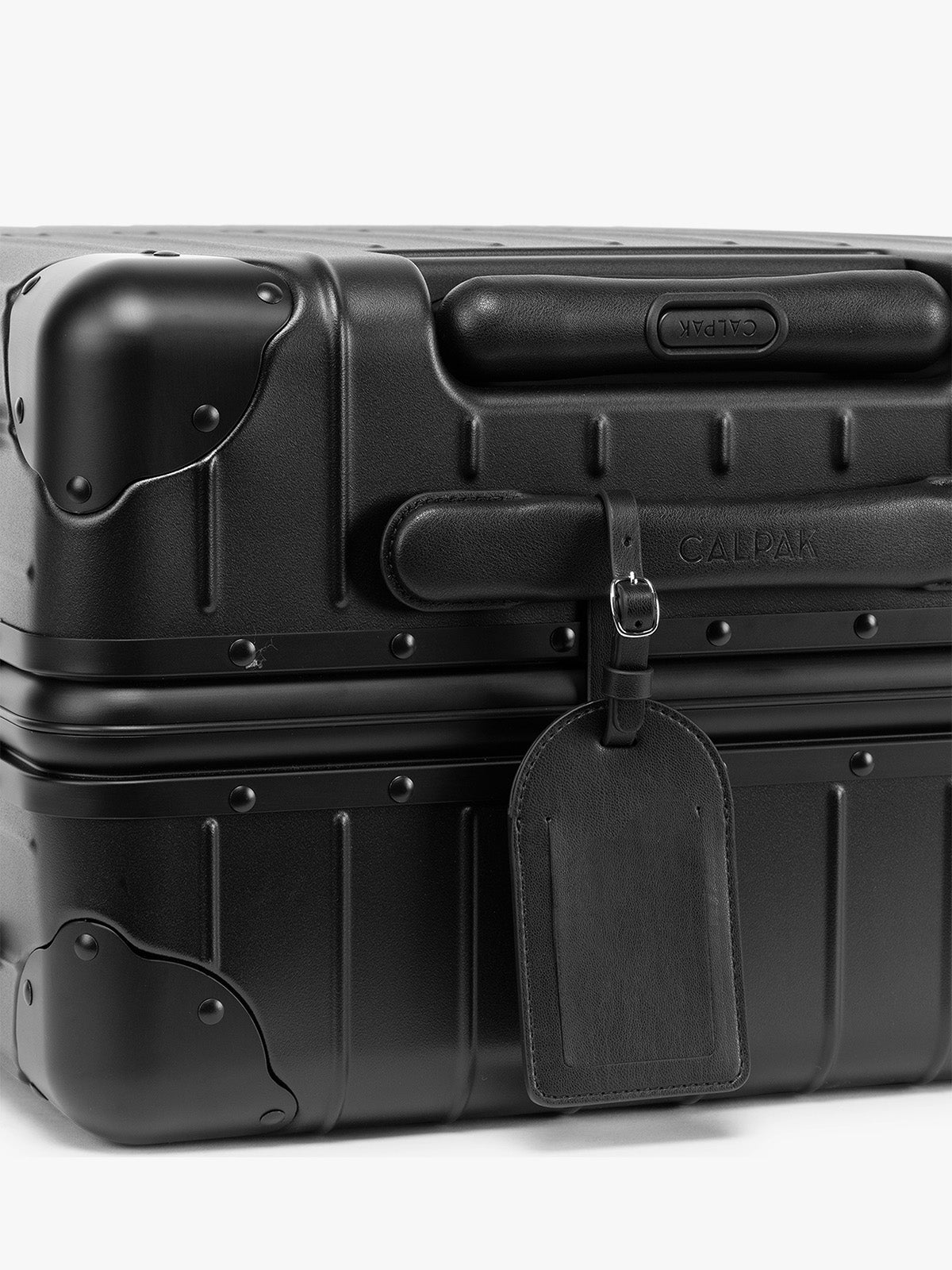 A close-up of a black hard-shell suitcase with a black CALPAK Luggage Tag attached, on a clean white background; PWTS2601-BLACK