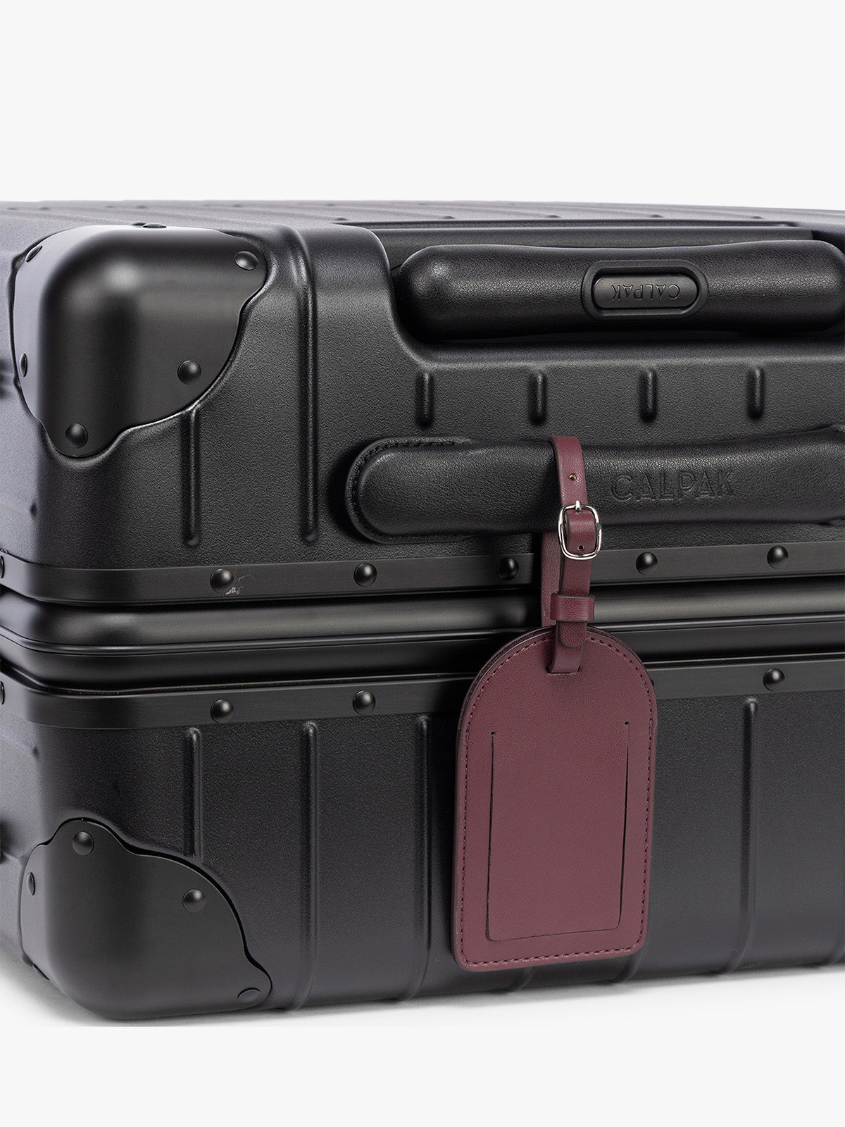 A close-up of a black hard-shell suitcase with a burgundy CALPAK Luggage Tag attached, on a clean white background; PWTS2601-DARK-CHERRY