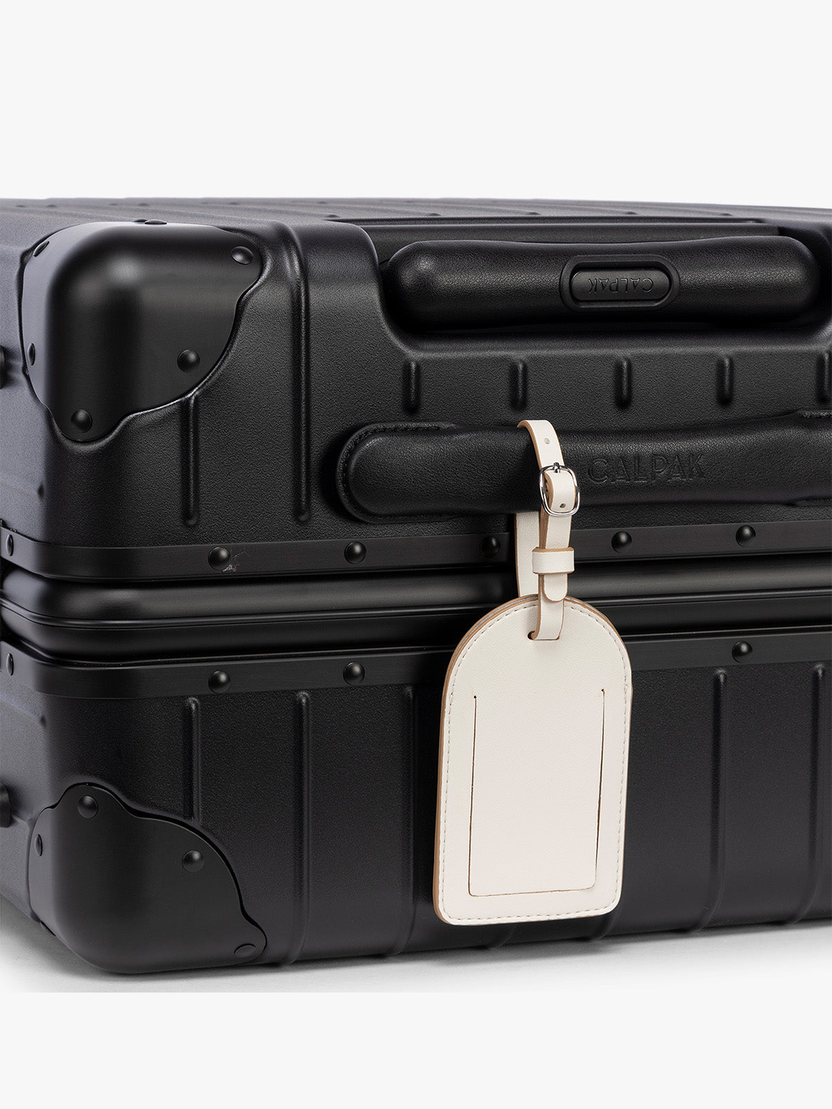 A close-up of a black hard-shell suitcase with a cream CALPAK Luggage Tag attached, on a clean white background; PWTS2601-VANILLA