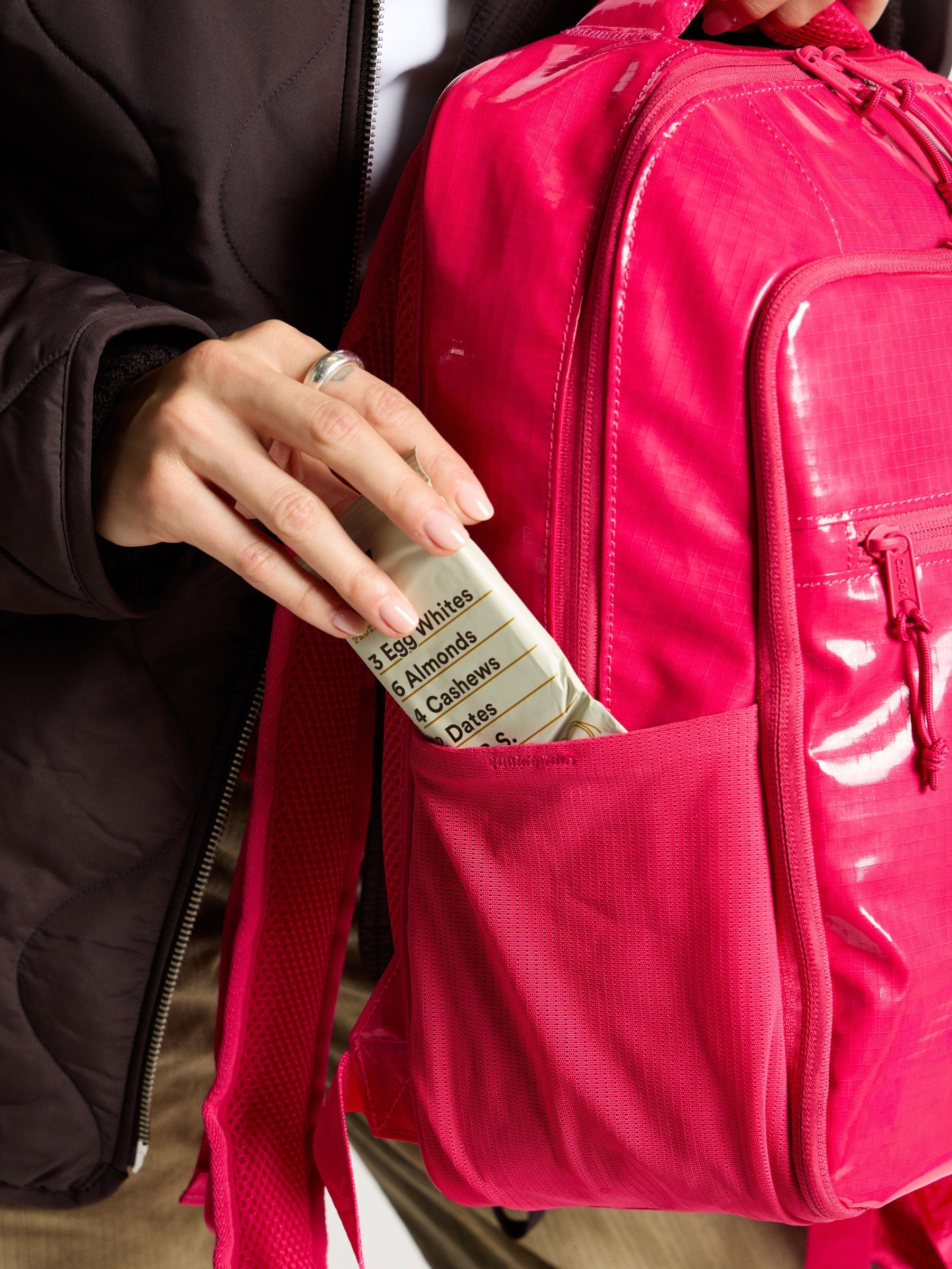 Model placing energy bar within the side pocket of CALPAK Terra Hydration Backpack in dragonfruit; BPT2201-DRAGONFRUIT