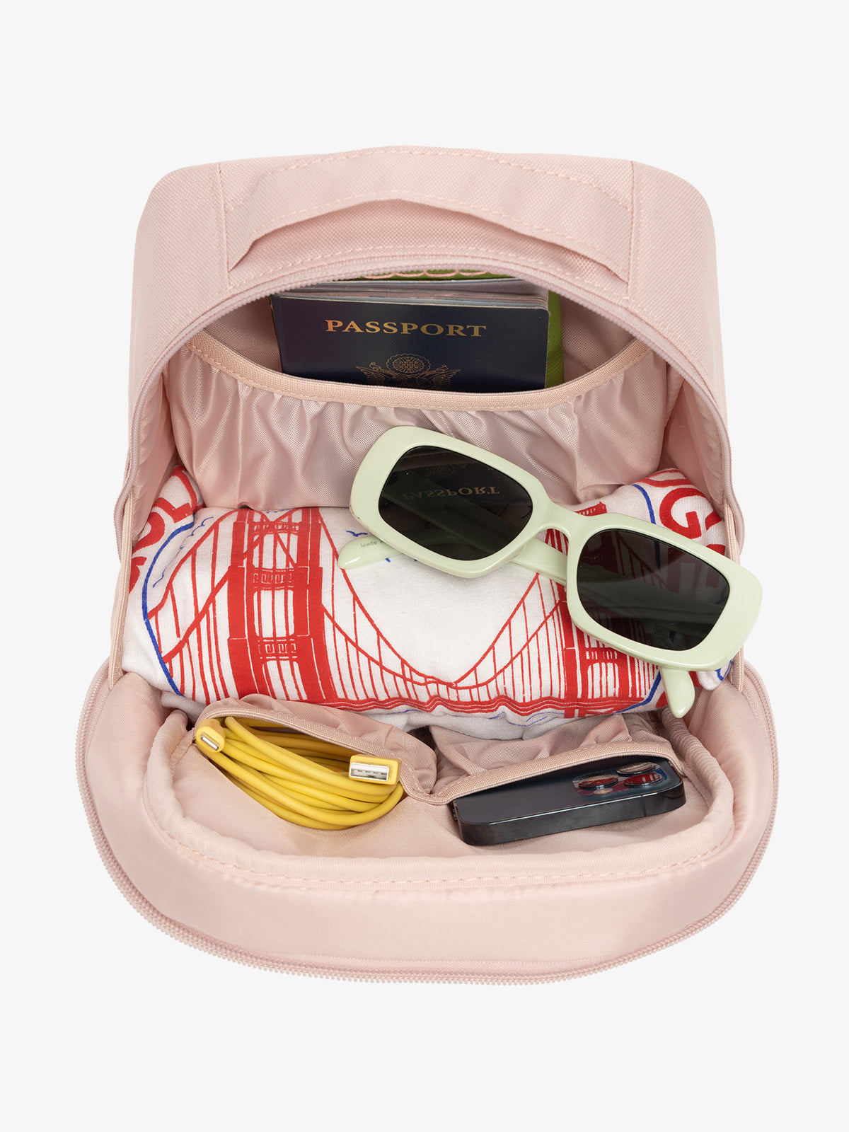 Interior view of pink sand CALPAK The Everything Cube with notebook and passport in large slip pocket, cords and phone in front slip pockets, and sunglasses and t-shirt in main compartment; ACE2401-PINK-SAND