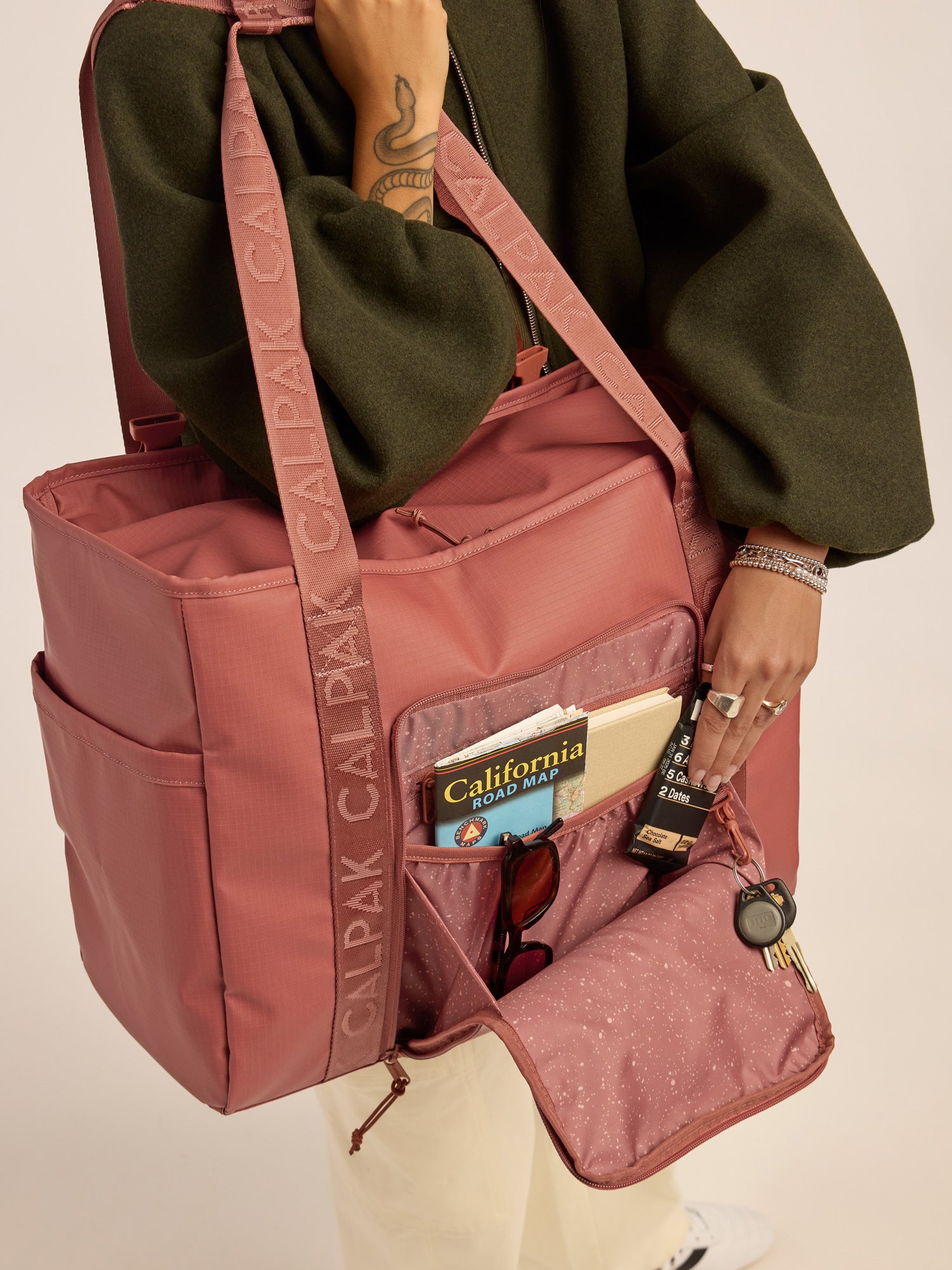 Model opening the front pocket of the CALPAK Terra 35L Water Resistant Zippered Tote Bag in red and orange Matte Clay; TTO2401-CLAY