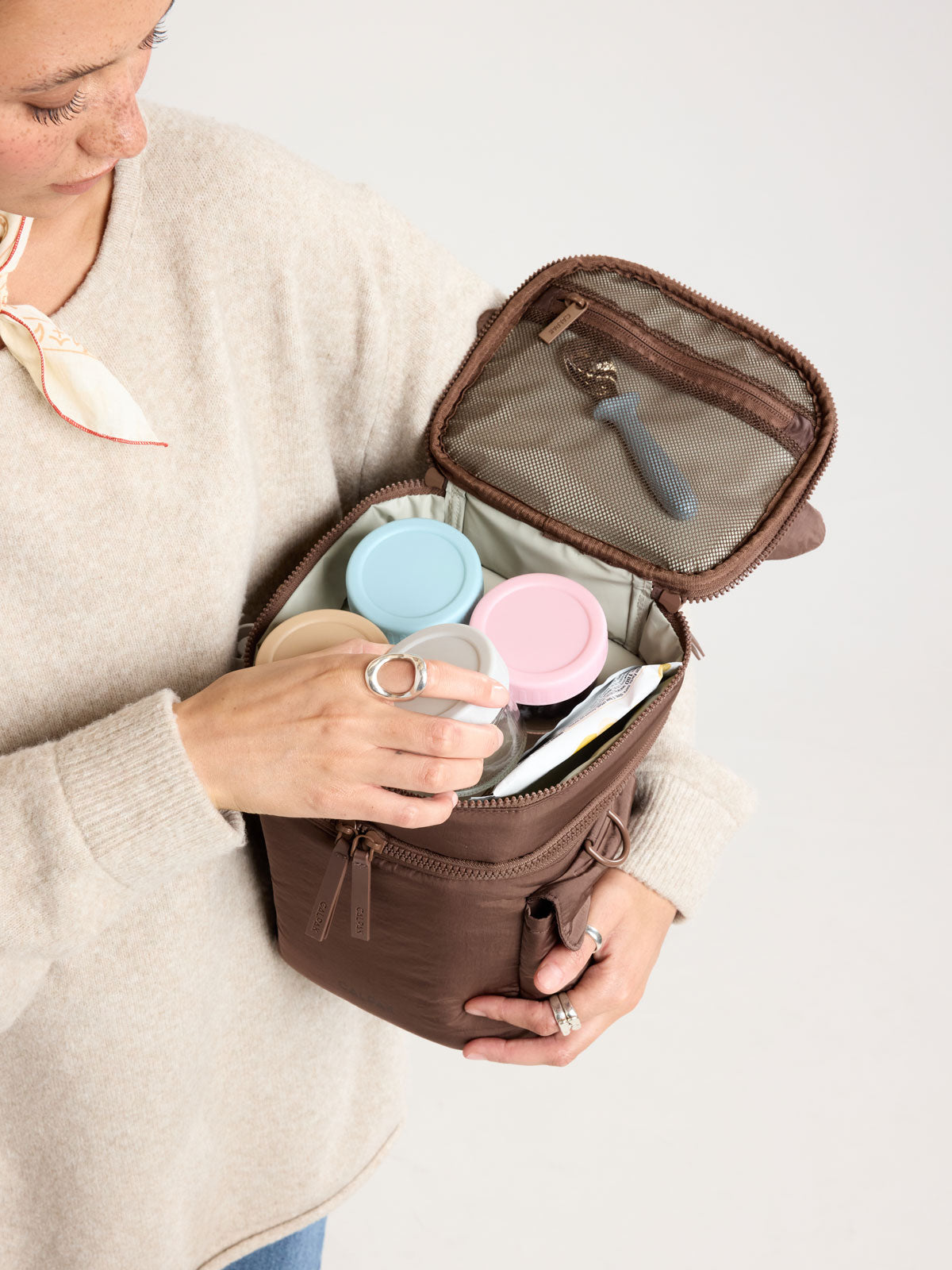 Woman adding items into CALPAK Insulated Baby Bottle Bag in brown Hazelnut; IBB2601-HAZELNUT