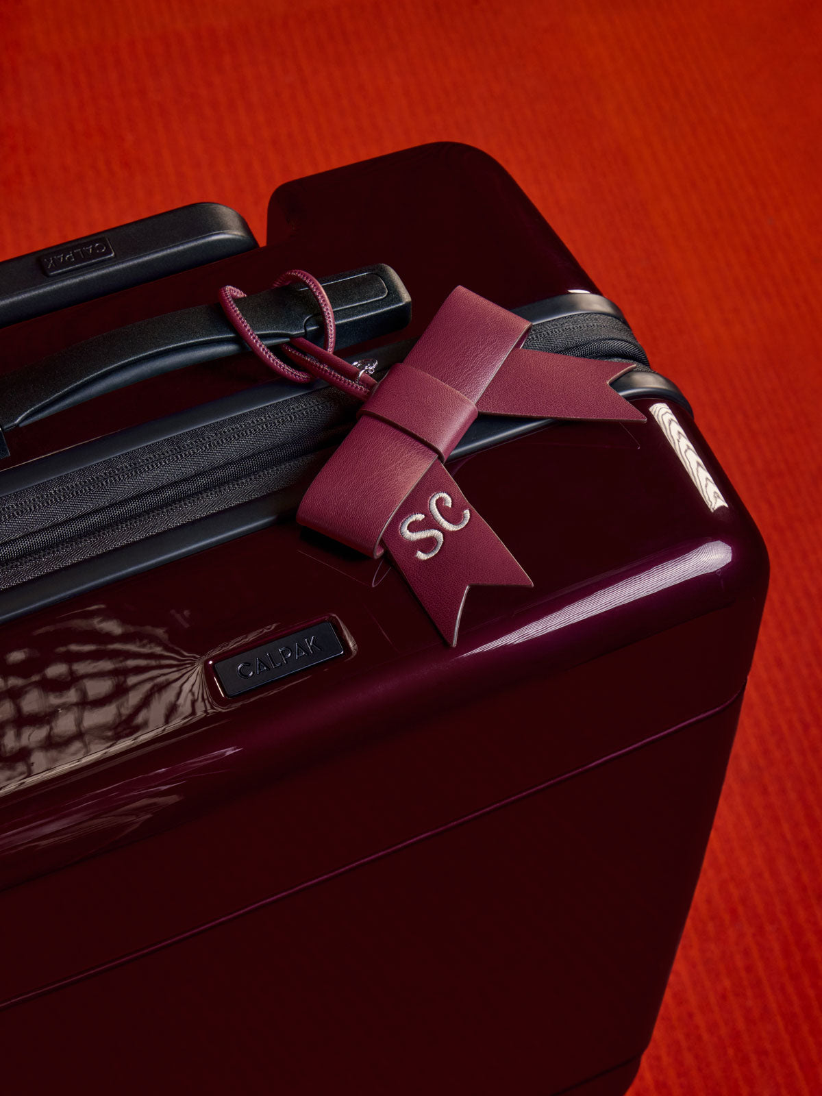 A close-up of a glossy dark burgundy suitcase with a matching burgundy CALPAK Personalized Bow Bag Charm in Dark Cherry tied around the handle, featuring a white embroidered 'SC' monogram and a textured orange-red background; BWBC2601-VANILLA