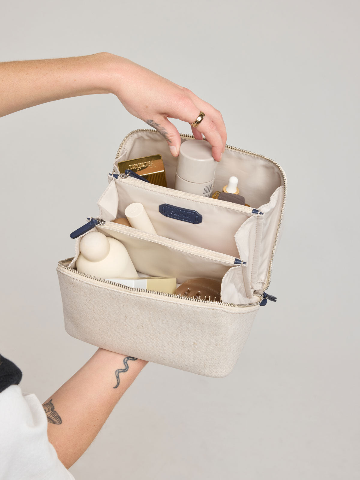 Woman putting items in CALPAK Colette Lay-Flat Canvas Toiletry Bag in beige canvas material and blue finishing; CLTB2601-NATURAL-NAVY
