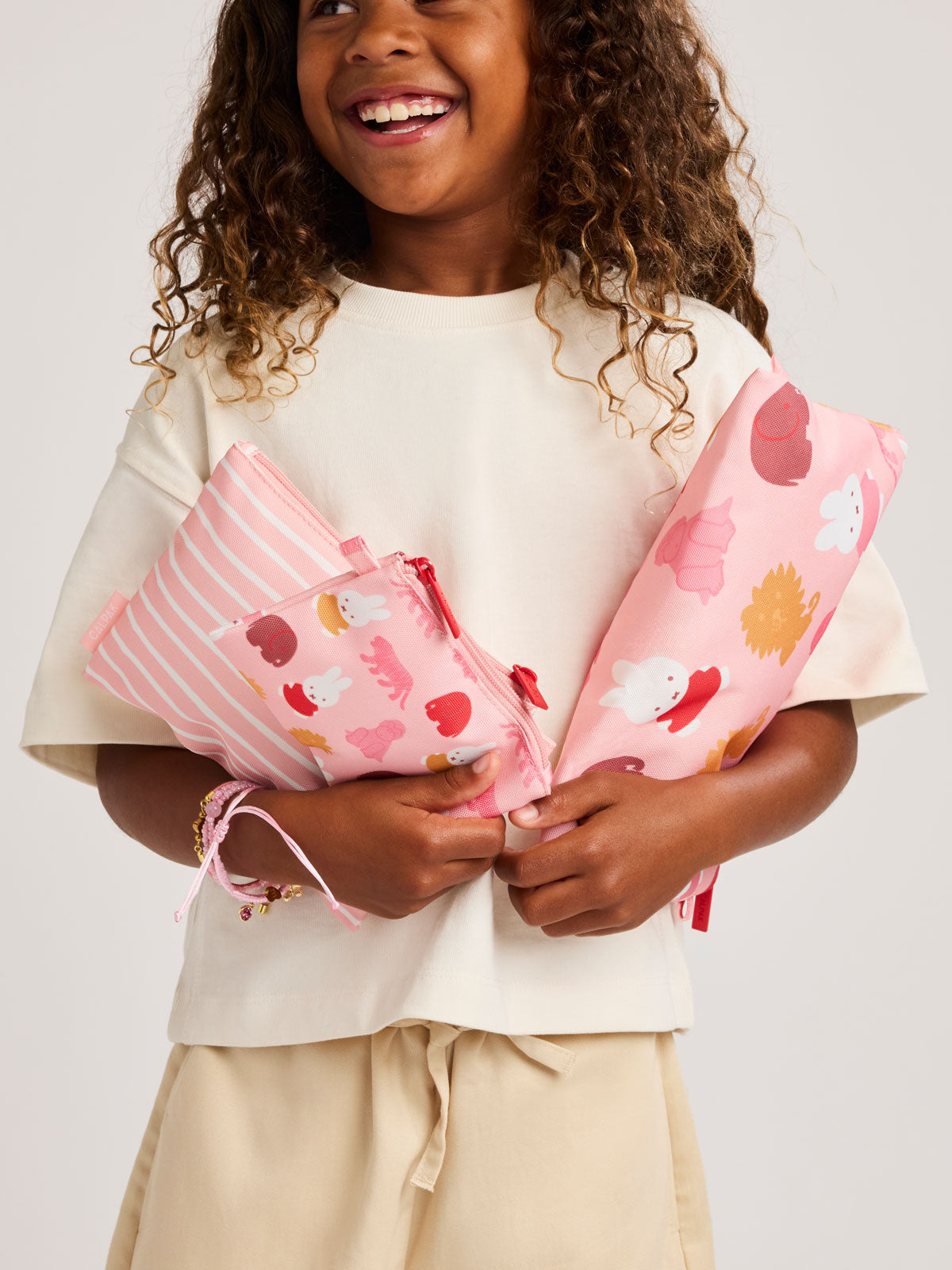 Child holding pink set of pink and white colorful children's pouches with Miffy and animal patterns for the CALPAK Compakt Zip Pouch Set on a white background; KZB2001-MIFFY-PINK-ZOO-TRIP